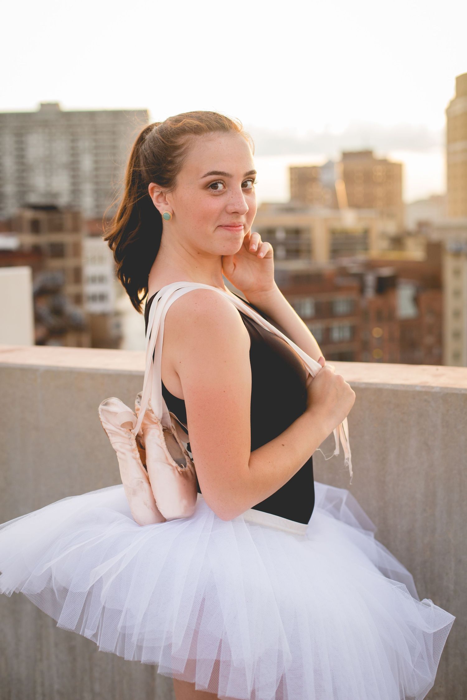 Young ballerina in white tutu holds pointe shoes on rooftop at sunset.