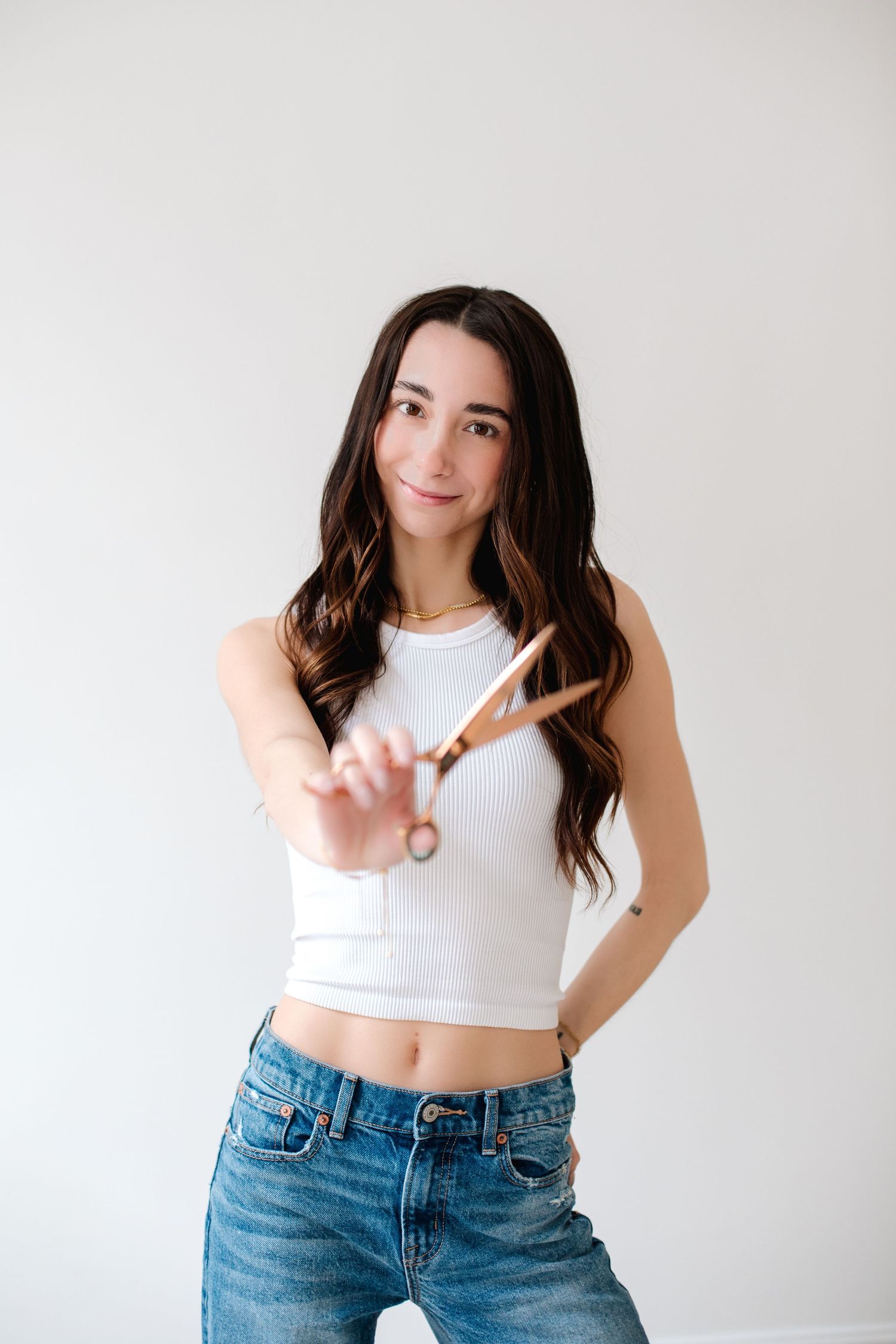 Brunette woman in white crop top and jeans holding gold scissors toward the camera.