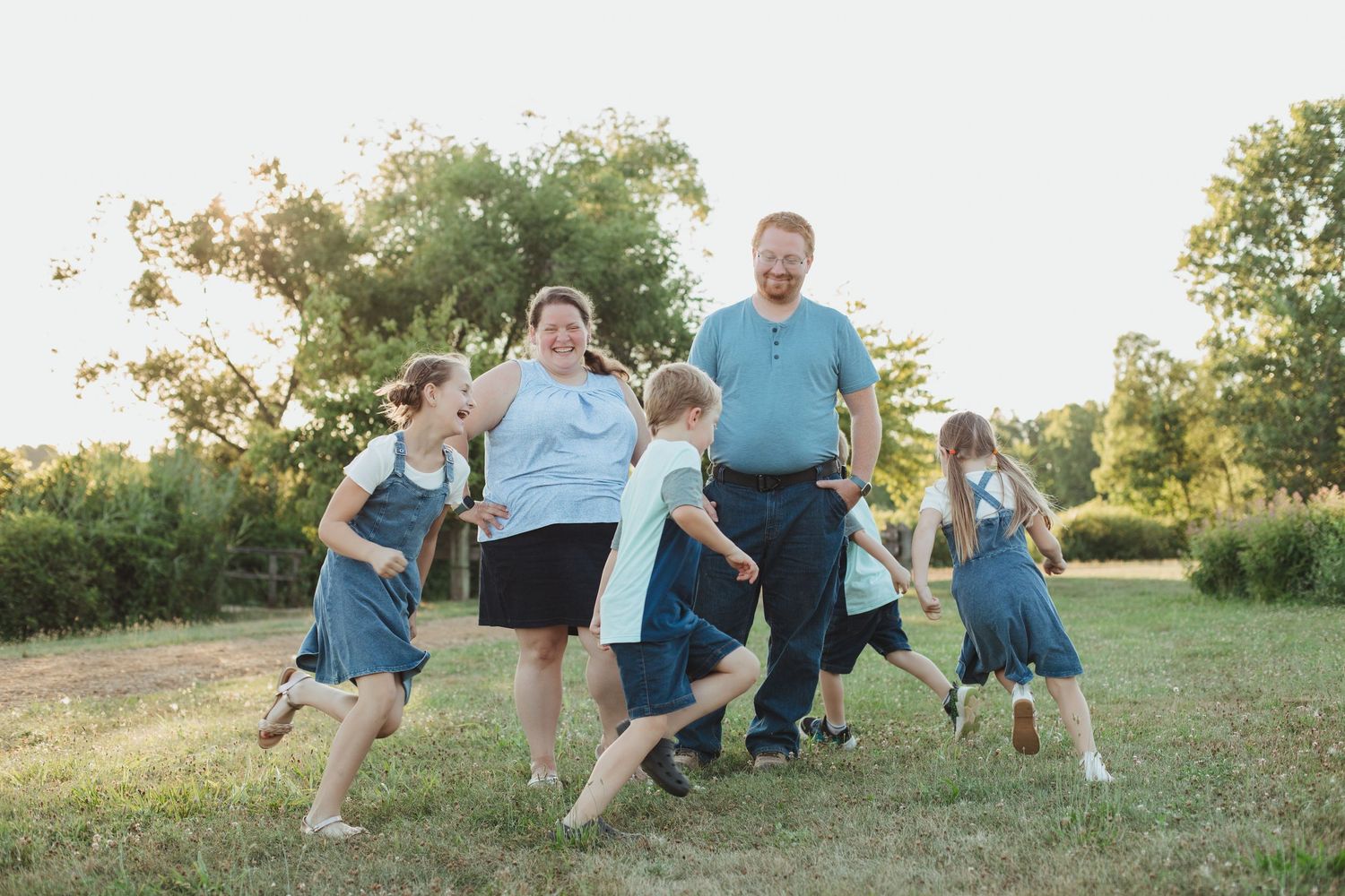 A joyful family plays together on a grassy field surrounded by trees in warm golden sunlight.