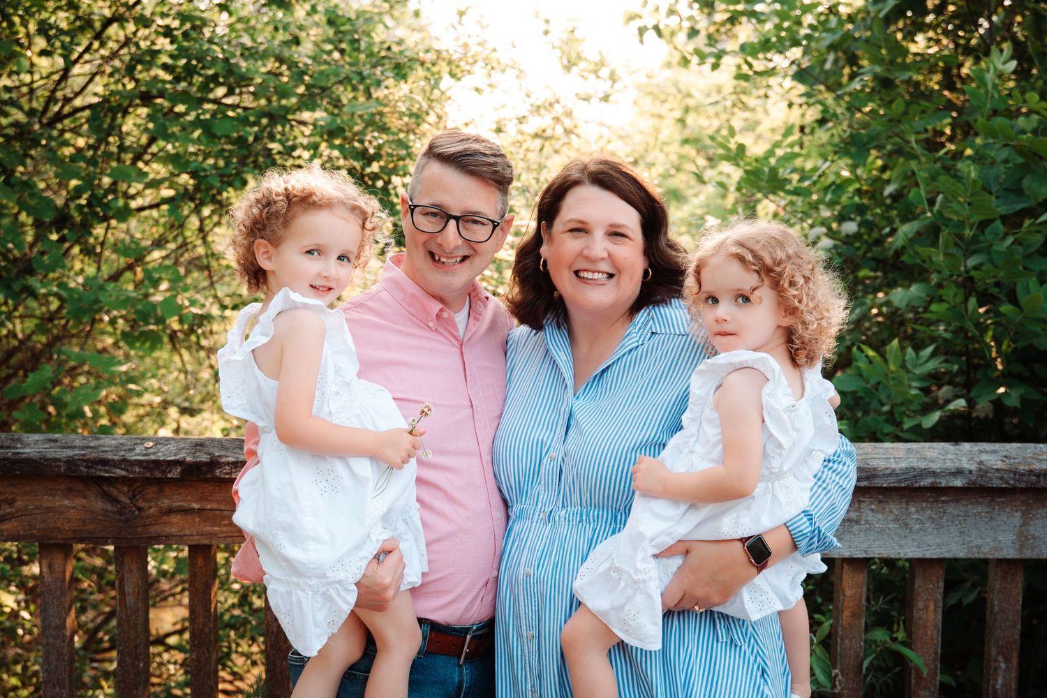 Happy family of four posing outdoors, parents holding twin girls in white dresses, smiling in a lush green setting.