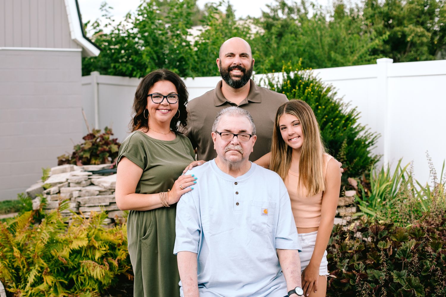 Family of four posing outdoors in a garden with white fence, including an elderly man seated in front.