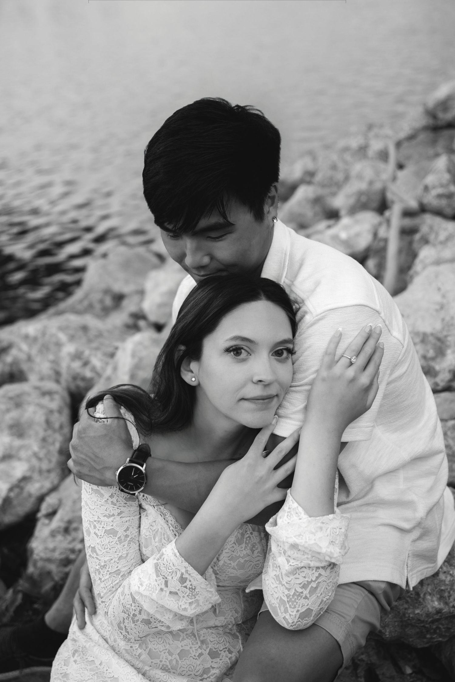 Black and white photo of a couple embracing near rocky shoreline, man kissing womans head while she looks at camera.
