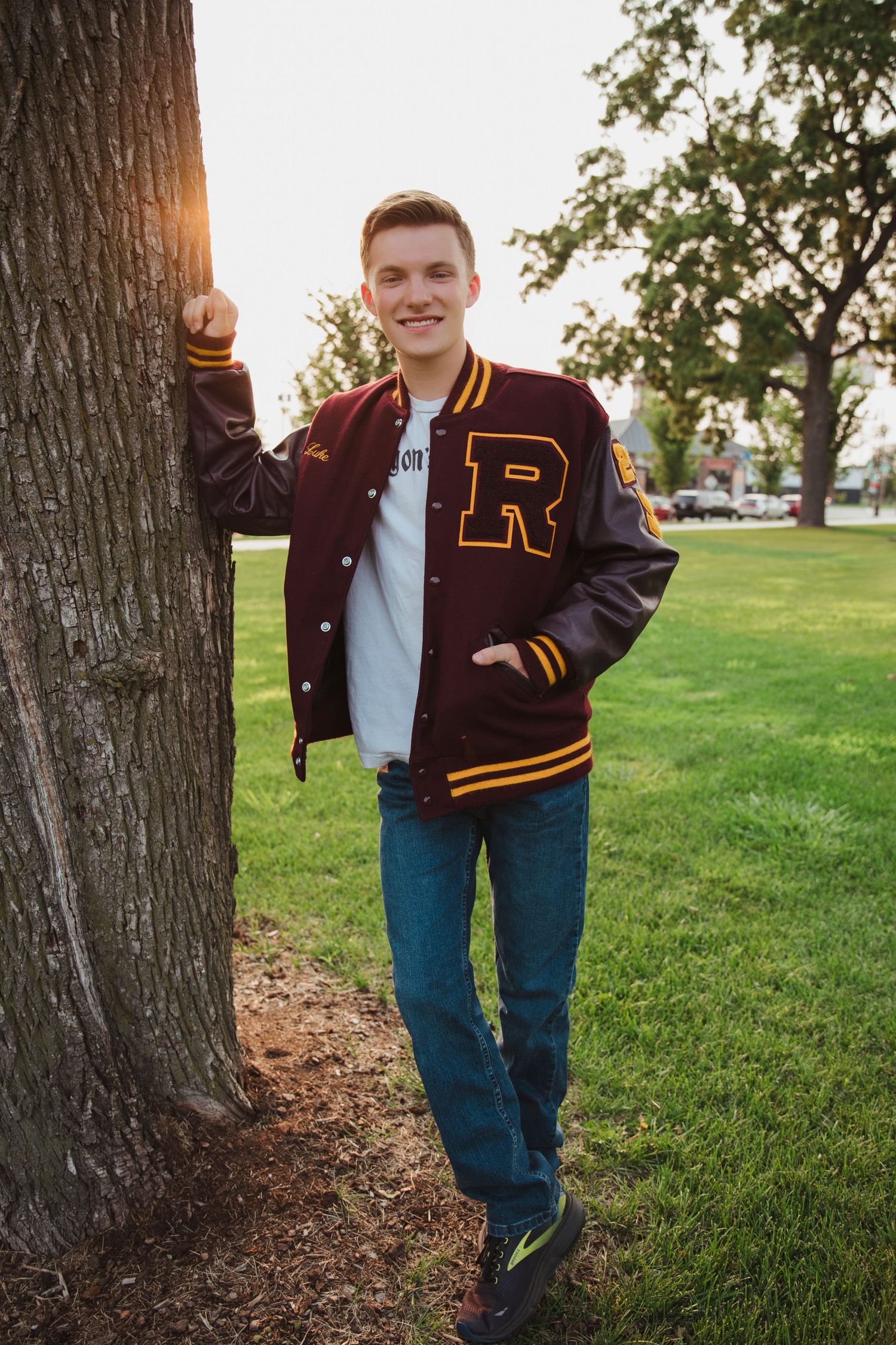 Young man in maroon R letterman jacket leaning against tree in sunny park setting.