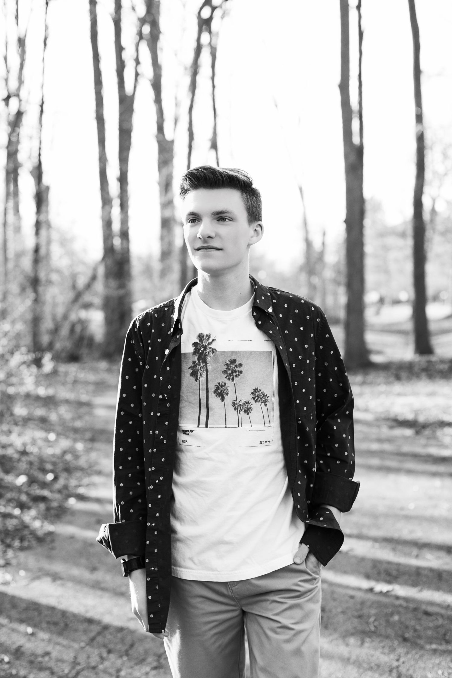Black and white portrait of young man in polka dot shirt standing among trees in park.