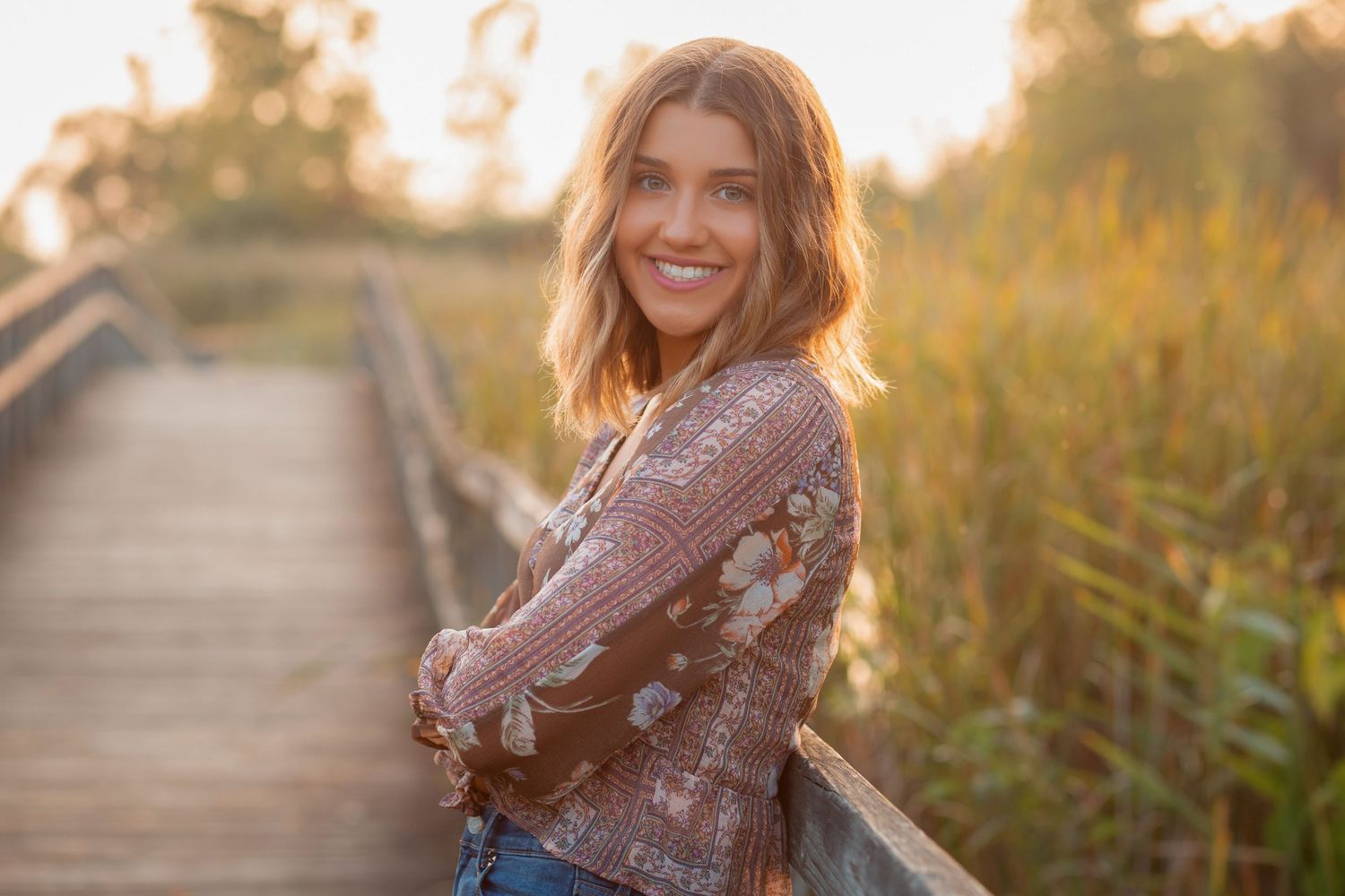 Smiling woman in floral jacket leaning on wooden bridge railing at golden hour.