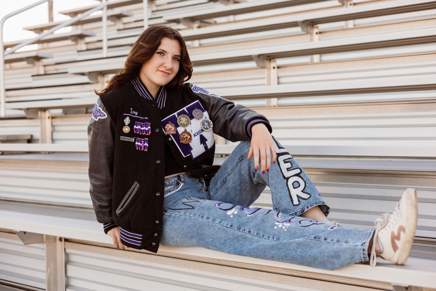 Teen girl in varsity letterman jacket sits relaxed on bleachers wearing jeans and sneakers.