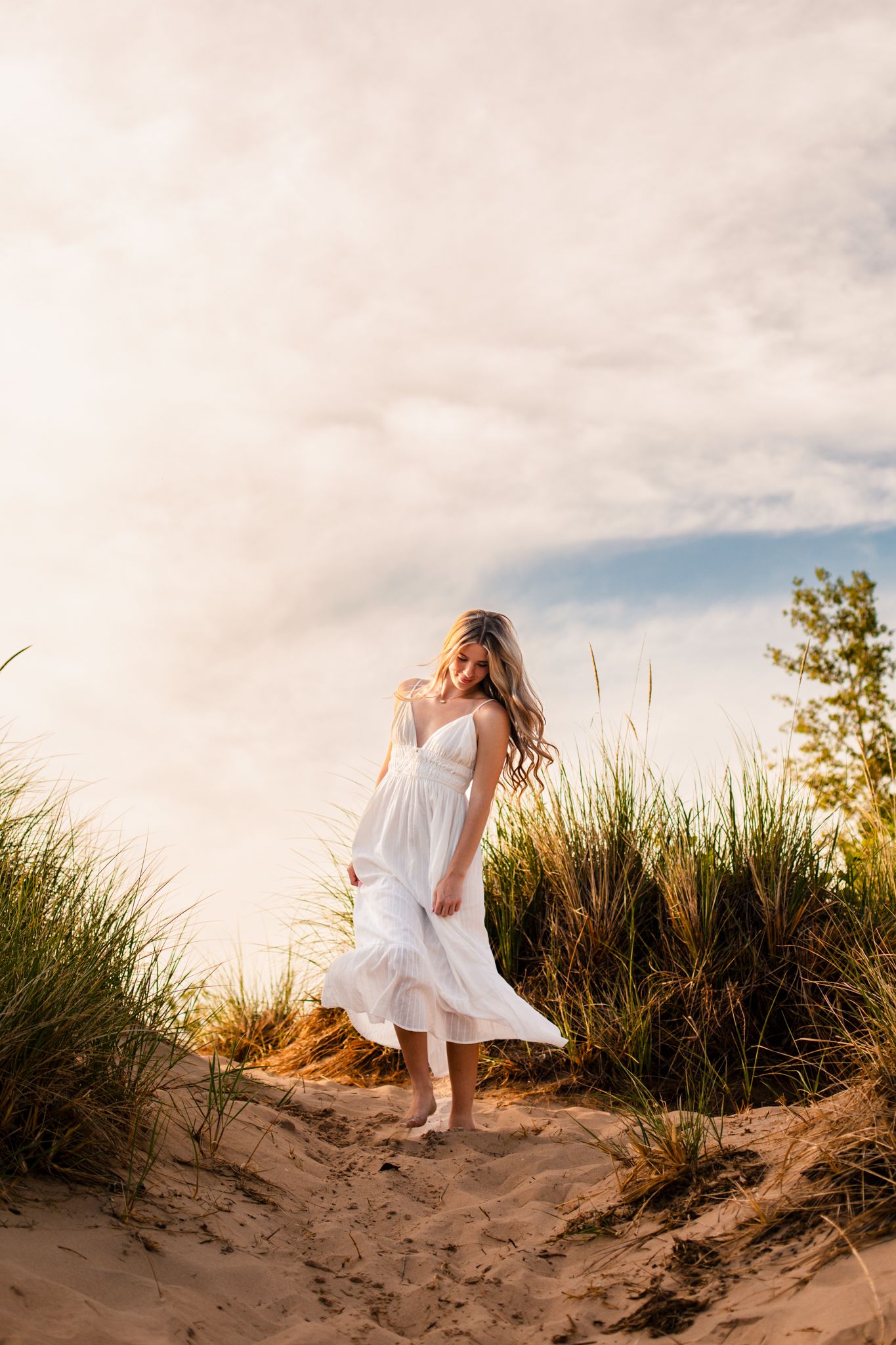 Young woman in white sundress standing on sand dunes surrounded by tall grass at golden hour.