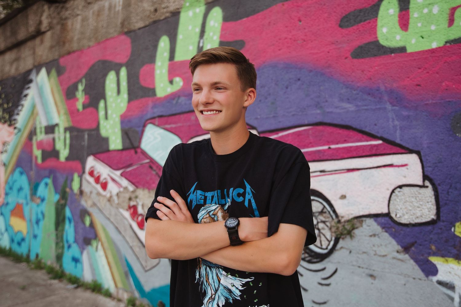 Smiling young man in black Metallica tee stands before colorful graffiti wall.