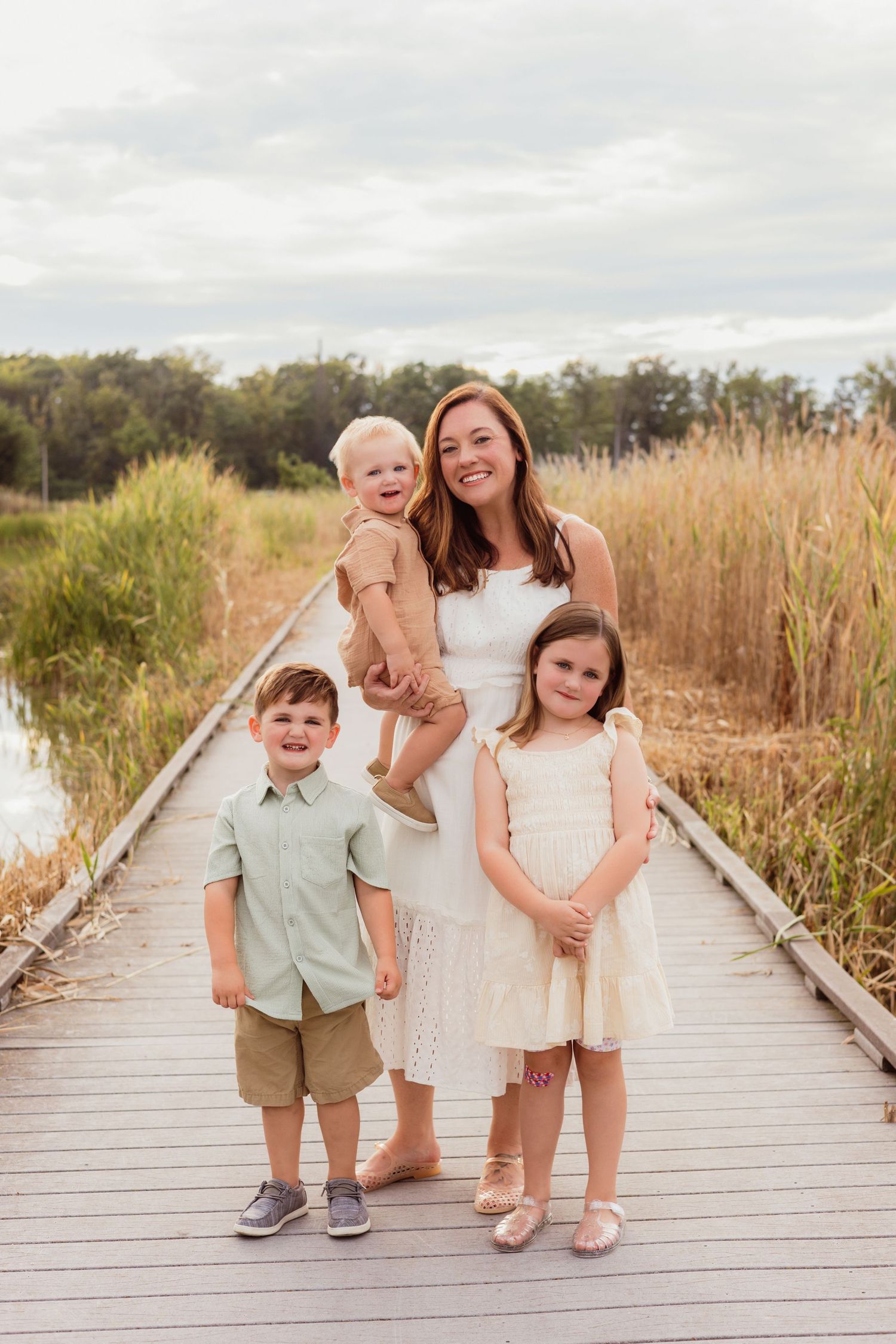 A smiling mother holds a baby while posing with two young children on a wooden boardwalk surrounded by tall grass.