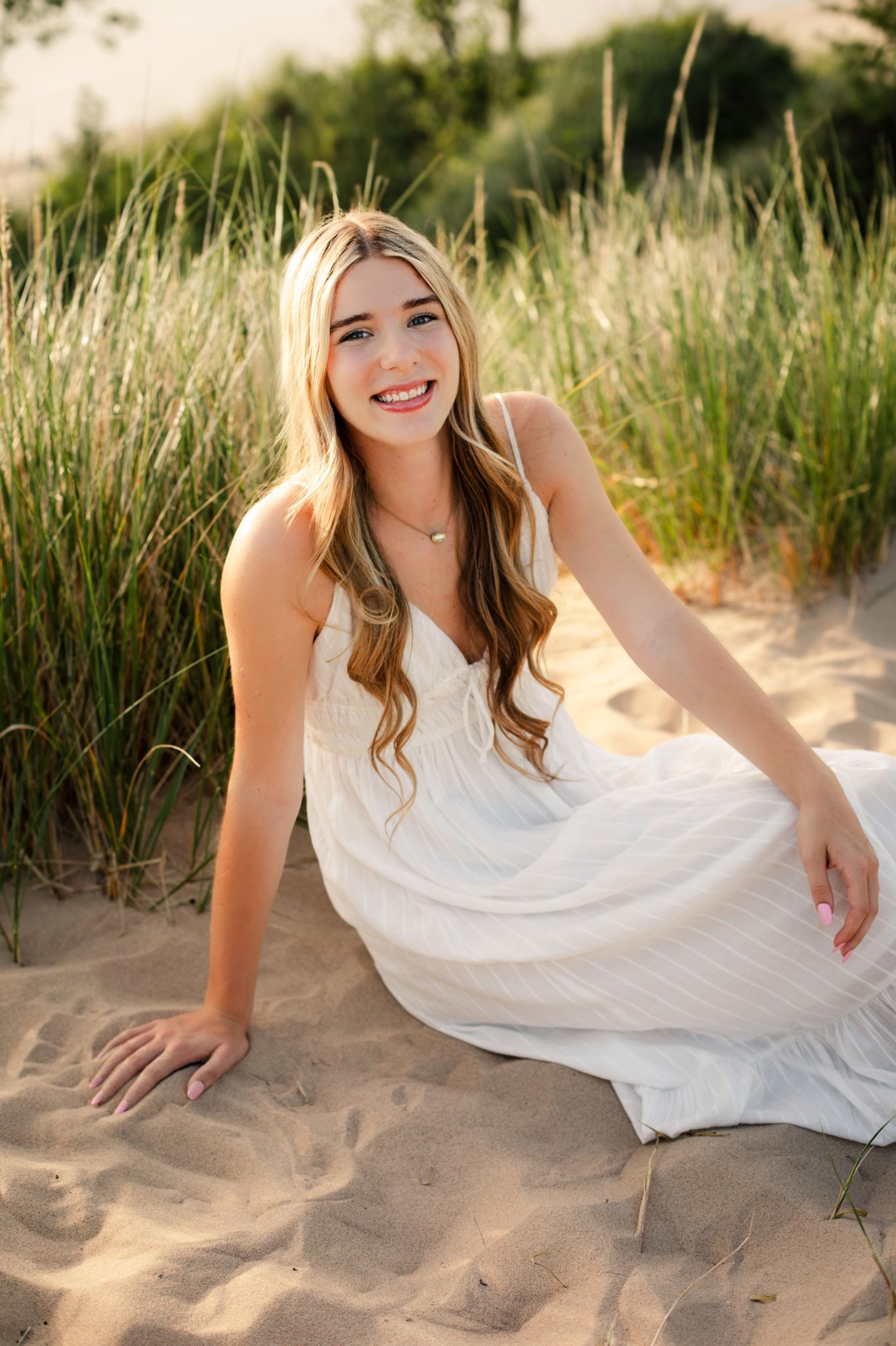 Young blonde woman in white sundress sitting on sandy beach dunes with tall grass, smiling outdoors.