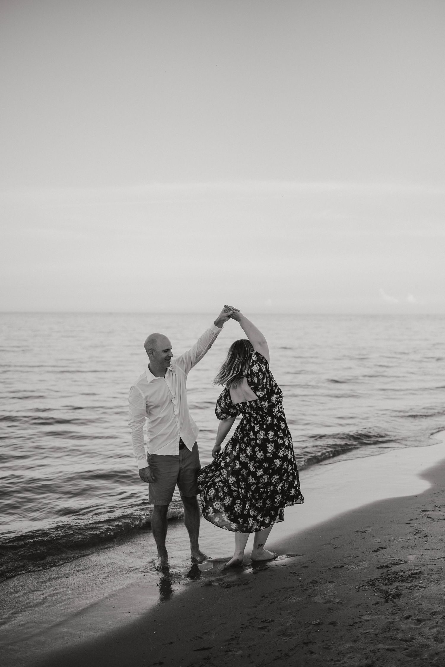 Black and white photo of a couple dancing barefoot together on a beach at the water's edge.