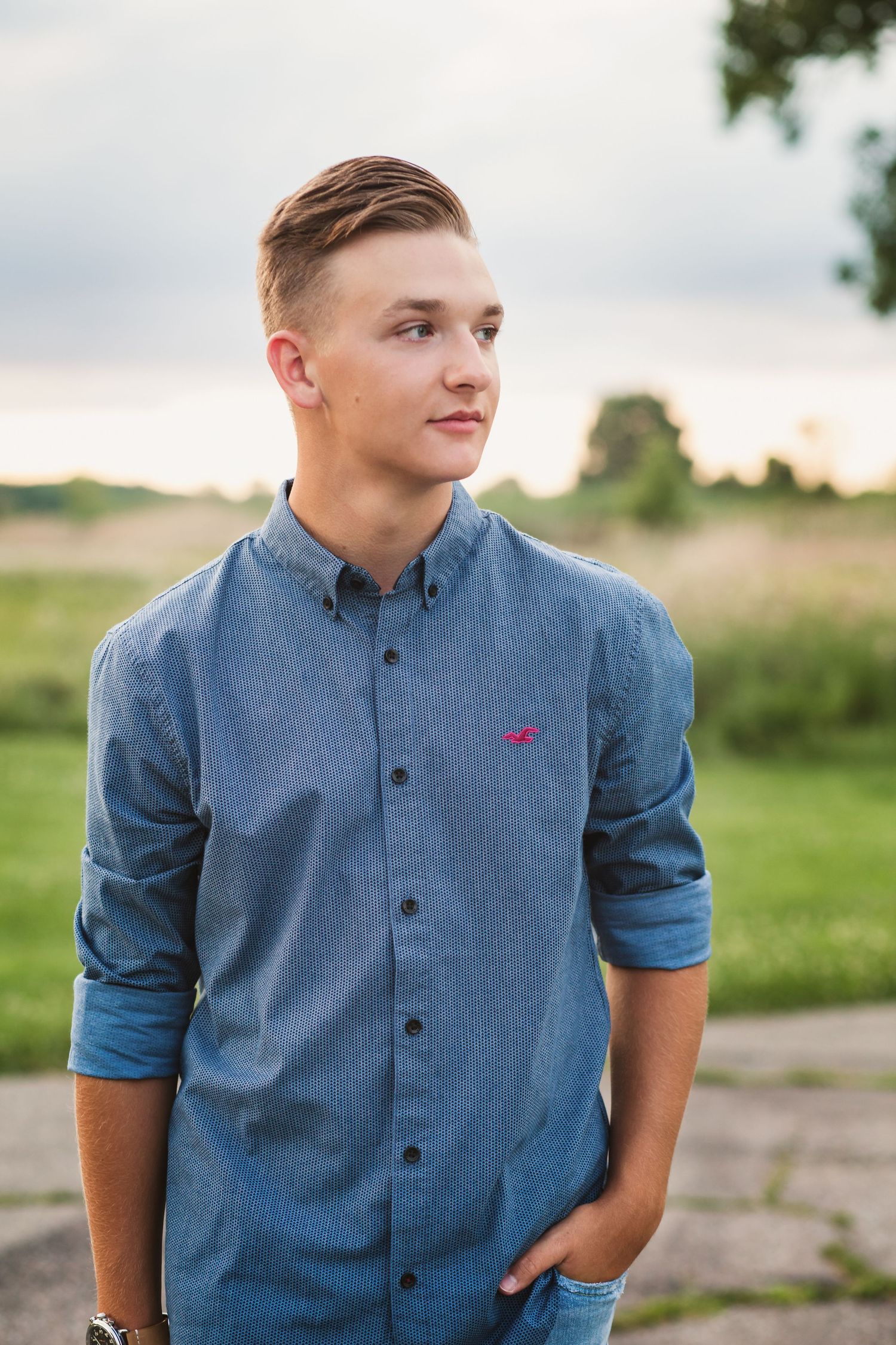 Young man in blue Hollister button-up shirt standing outdoors in a field at sunset, looking thoughtfully to the side.