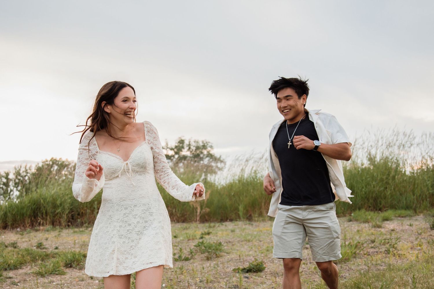 A happy couple runs hand in hand through a grassy field, the woman wearing a white lace dress.