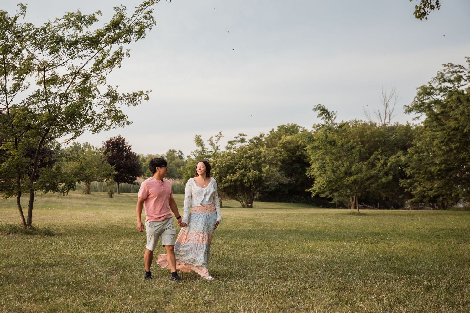 Couple walking hand in hand through a lush green park on a warm summer evening.