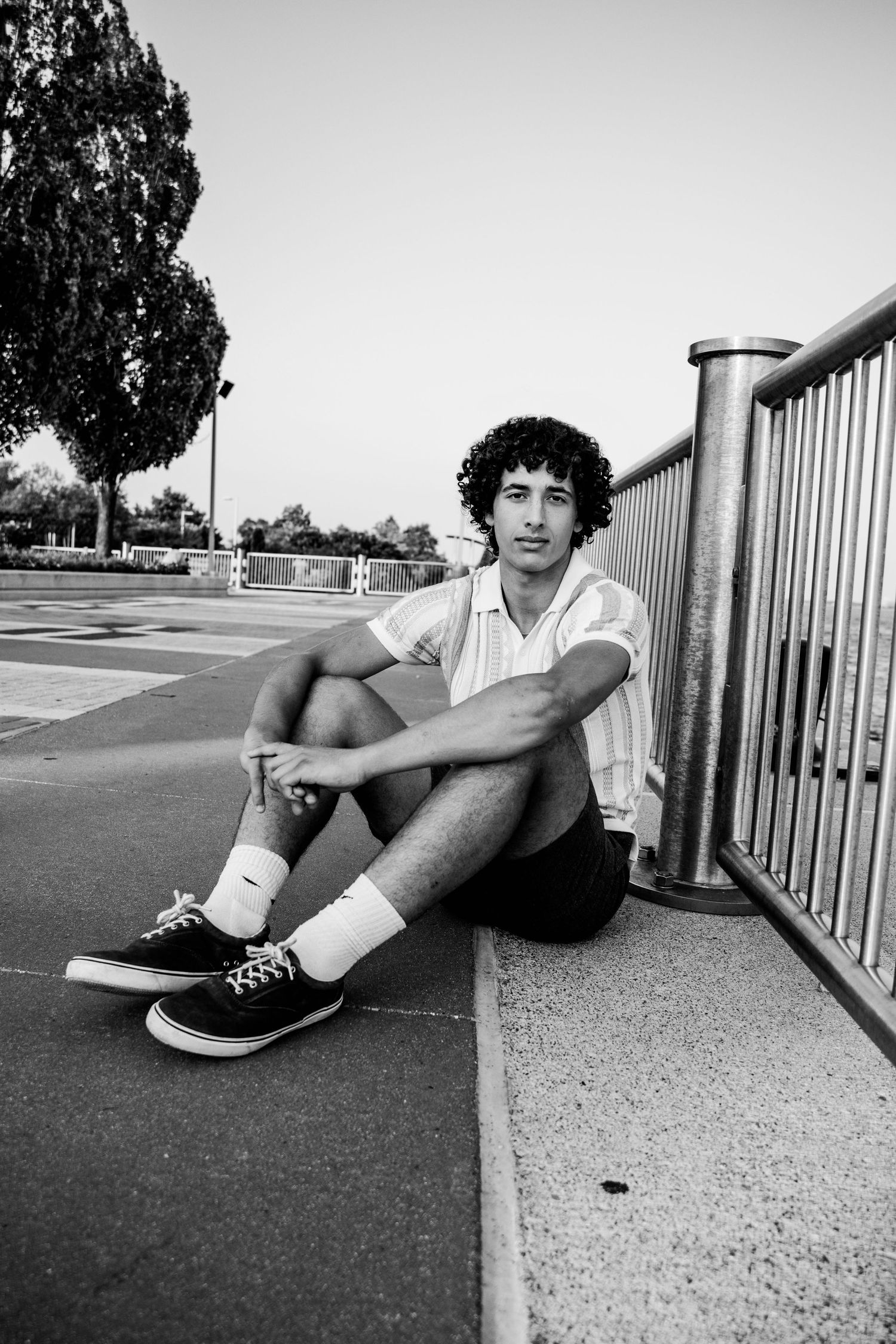 Black and white photo of a young man with curly hair sitting on pavement wearing shorts, white socks, and Vans sneakers.