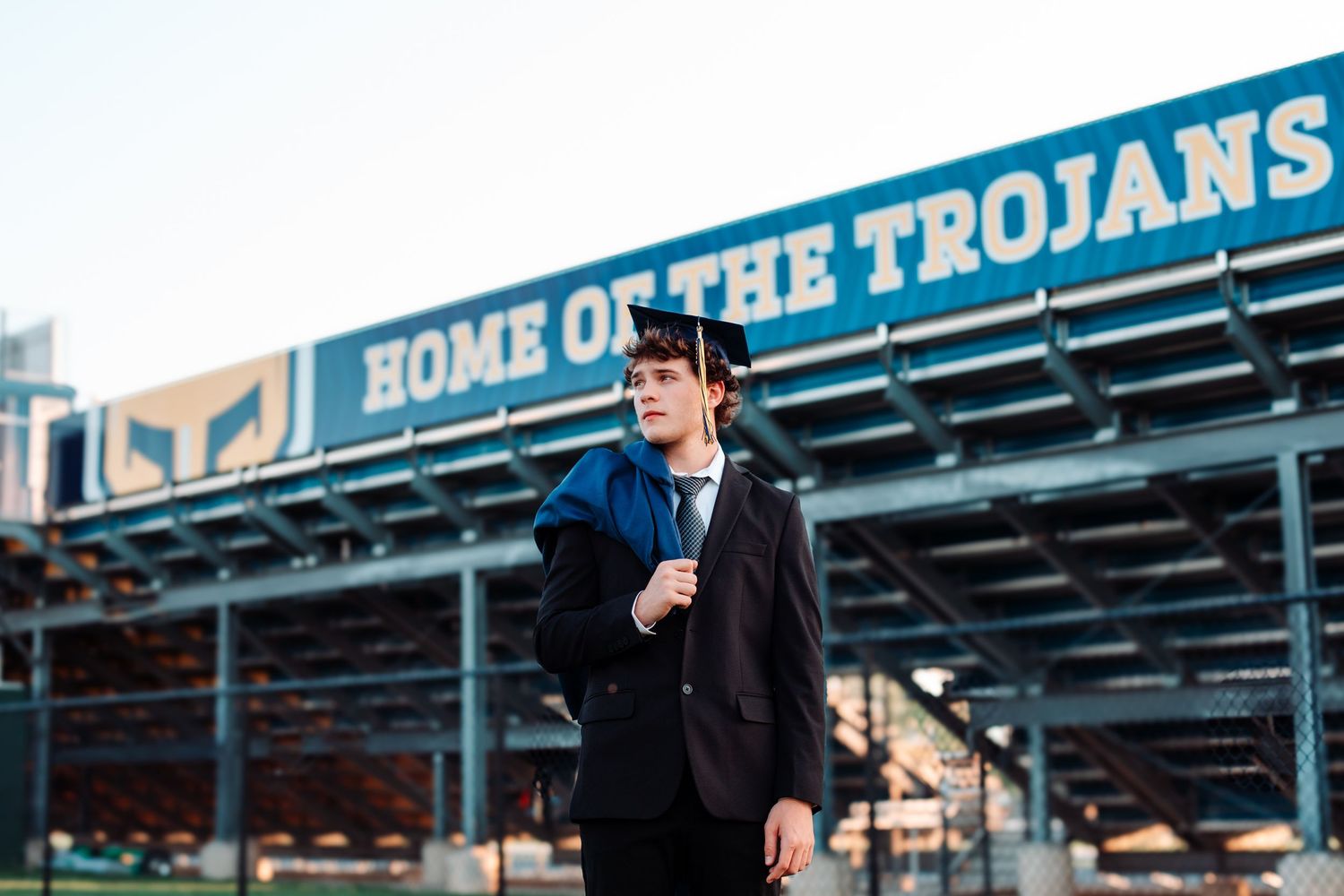 Graduate in cap and gown stands confidently outside a stadium marked Home of the Trojans.