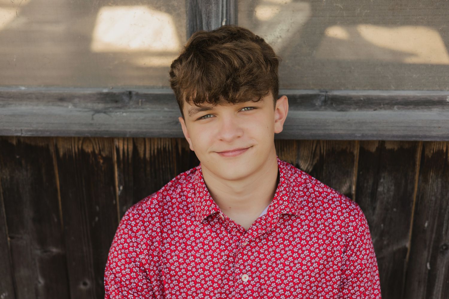 Smiling teenage boy with brown hair wearing a red floral button-up shirt, posing outdoors near a wooden wall.