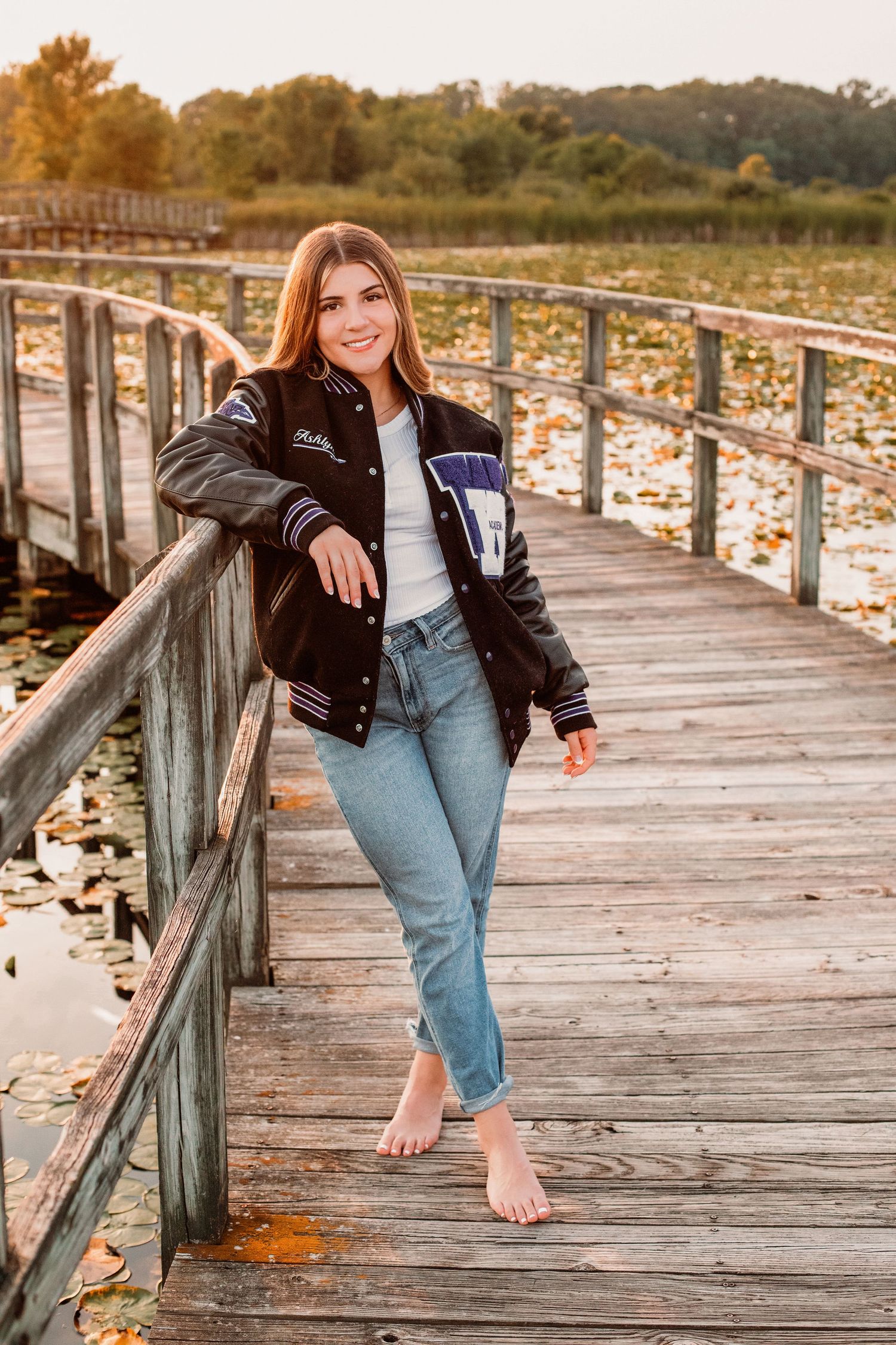 Young woman in varsity jacket and jeans poses barefoot on wooden dock over lily pad-covered lake at sunset.