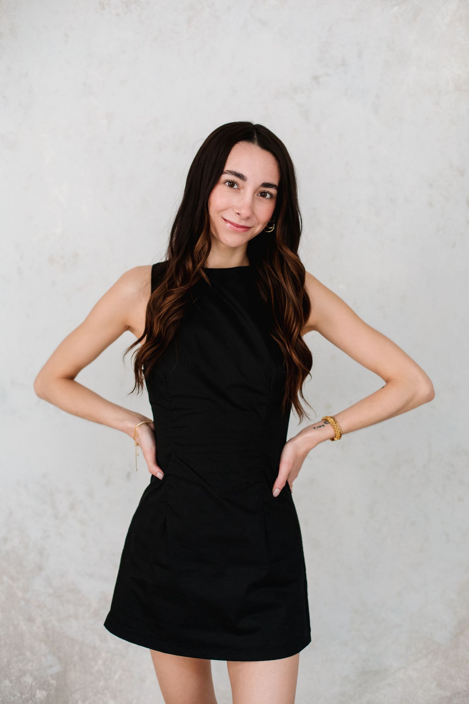 Young brunette woman in black mini dress posing with hands on hips against textured white wall.