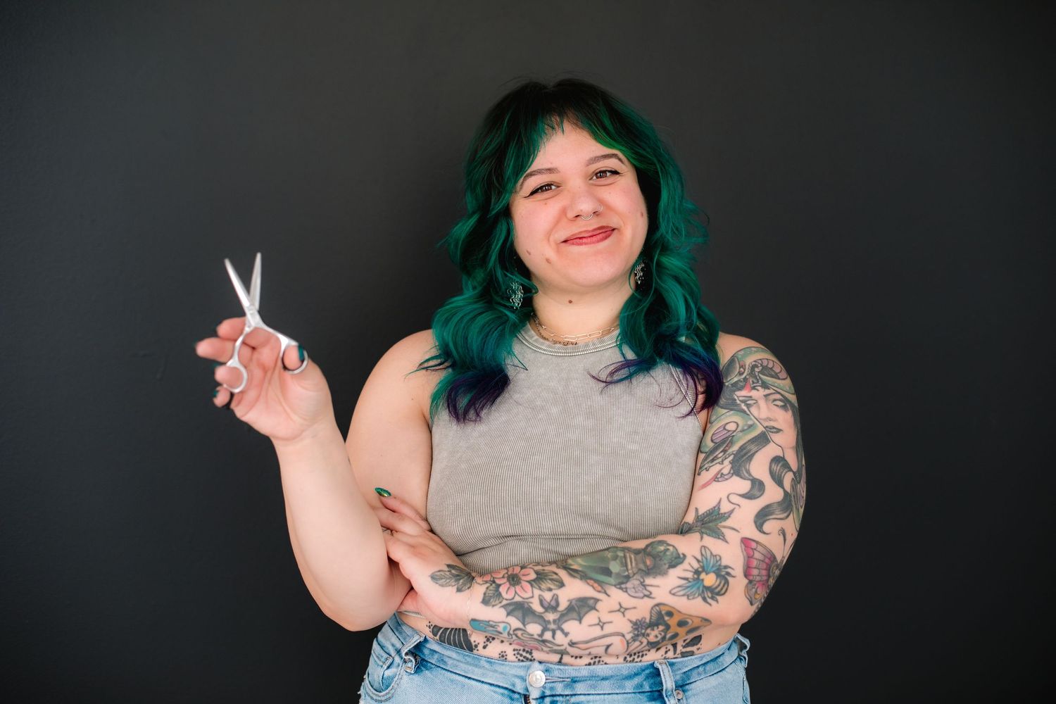 Smiling woman with teal hair and tattoos holding scissors against dark background.