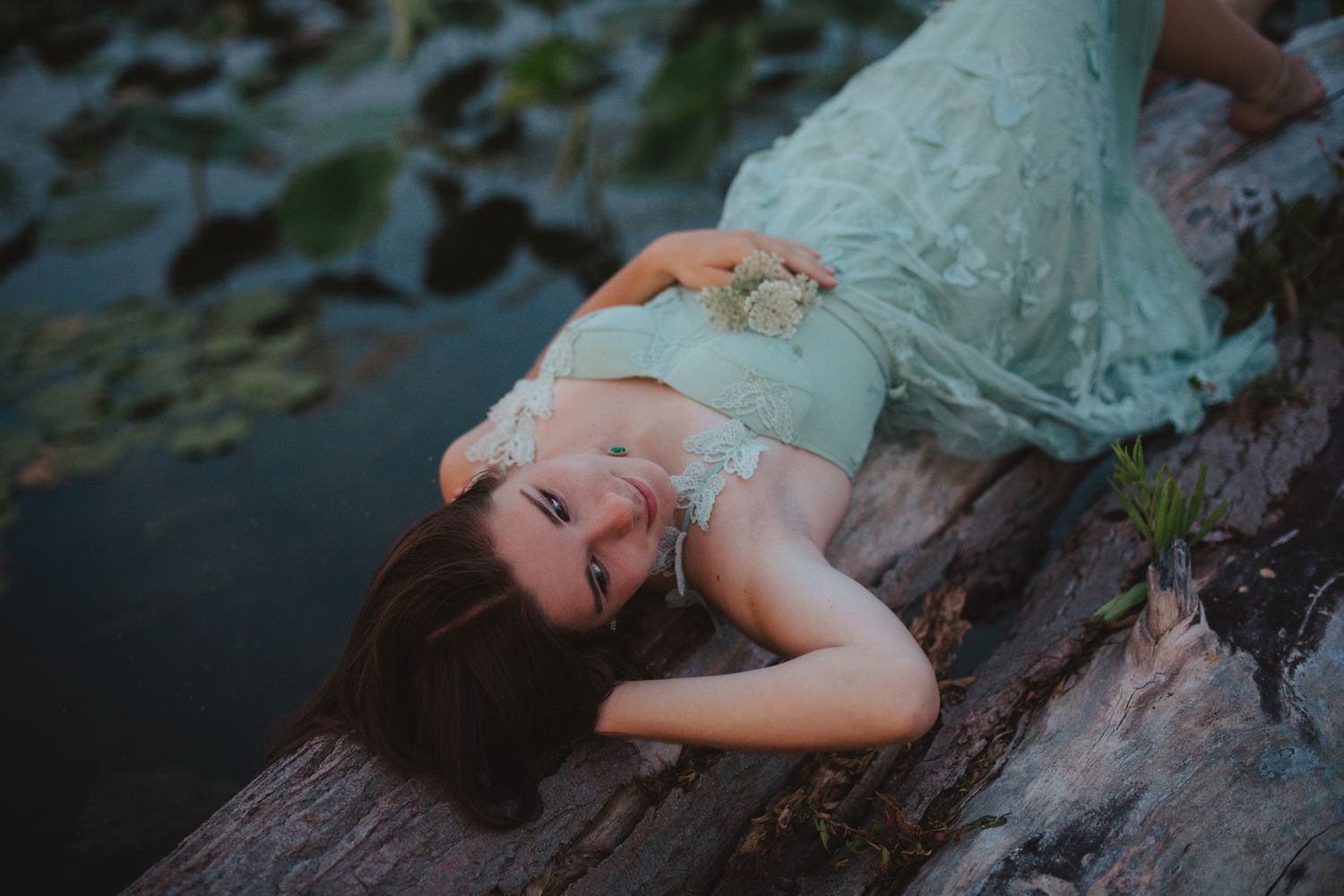 Young woman in mint lace dress lying on a log by a tranquil pond surrounded by lush greenery.