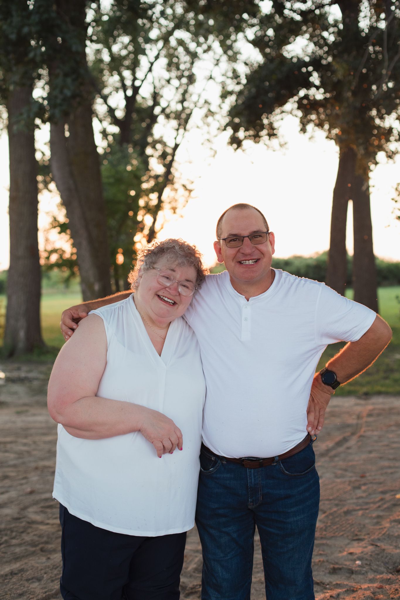 Smiling couple in matching white tops posing together outdoors at golden hour near trees.