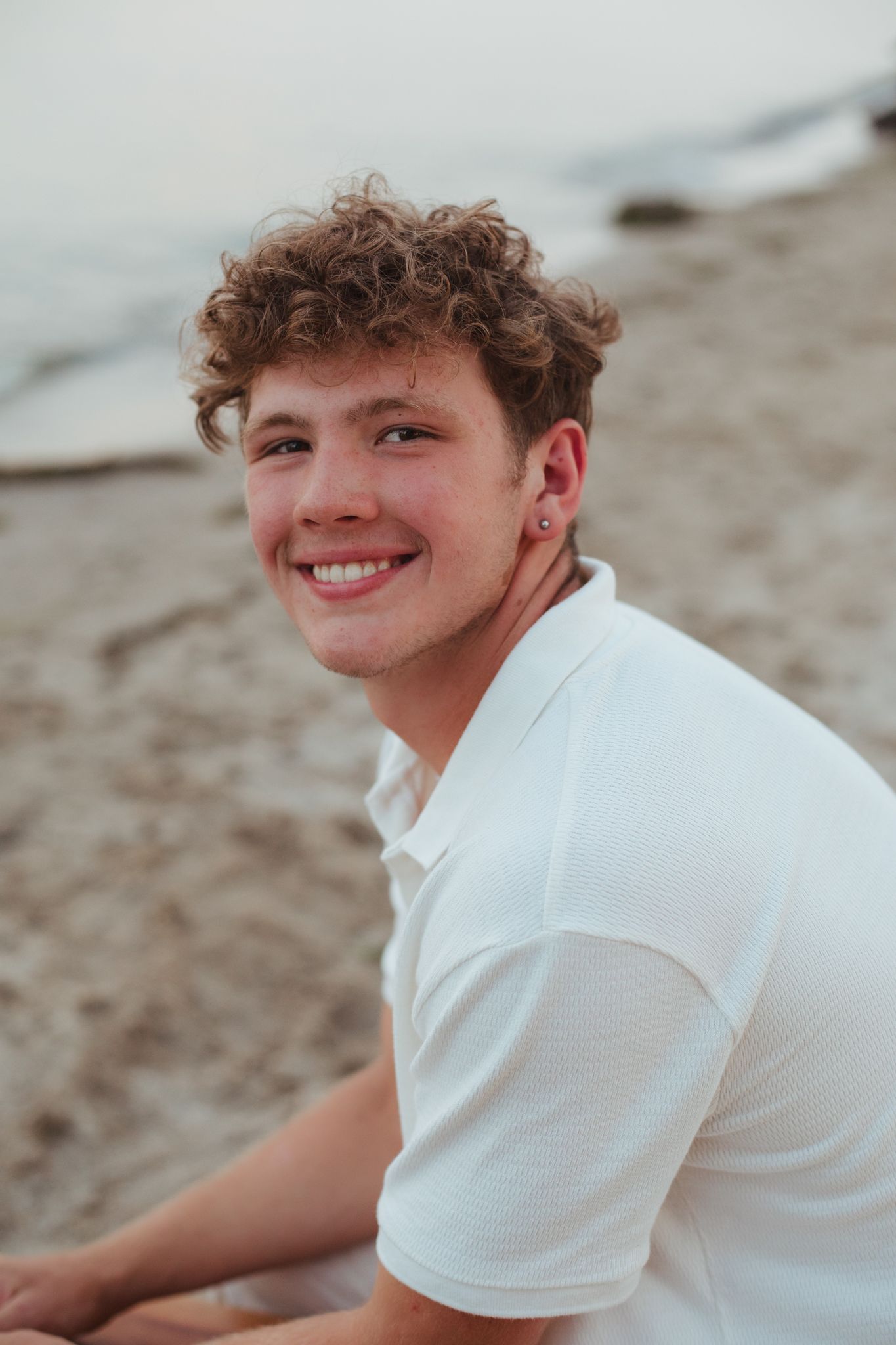 Smiling young man with curly hair wearing a white polo shirt sitting on a sandy beach near the water.