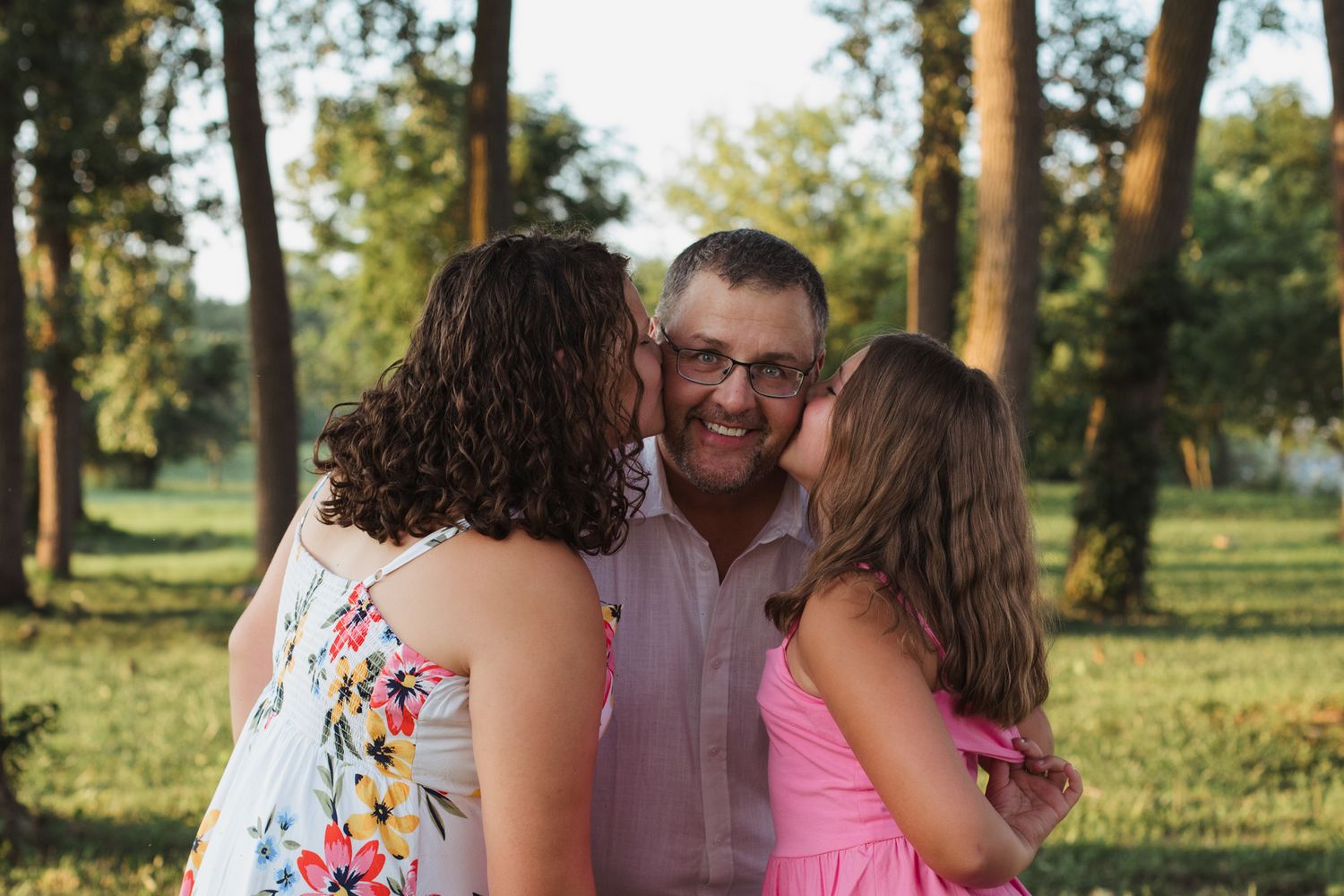 A smiling man receives kisses on both cheeks from two females in a sunny green park setting.