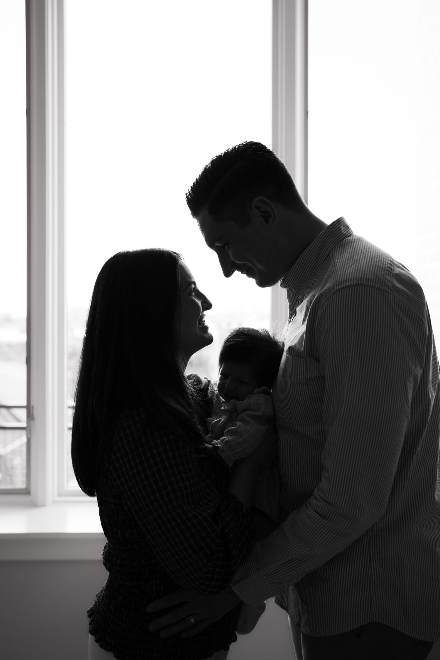 Black and white silhouette of a couple holding their newborn baby by a bright window.