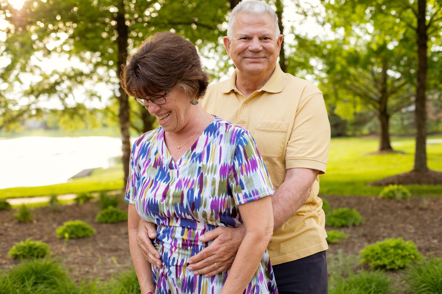 Older couple smiling together outdoors in a lush green park setting on a warm sunny day.