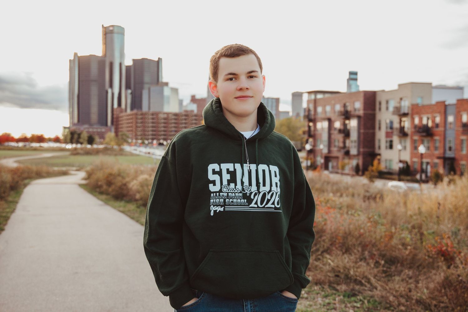 High school senior in dark hoodie poses on pathway with Detroit Renaissance Center skyline in background.
