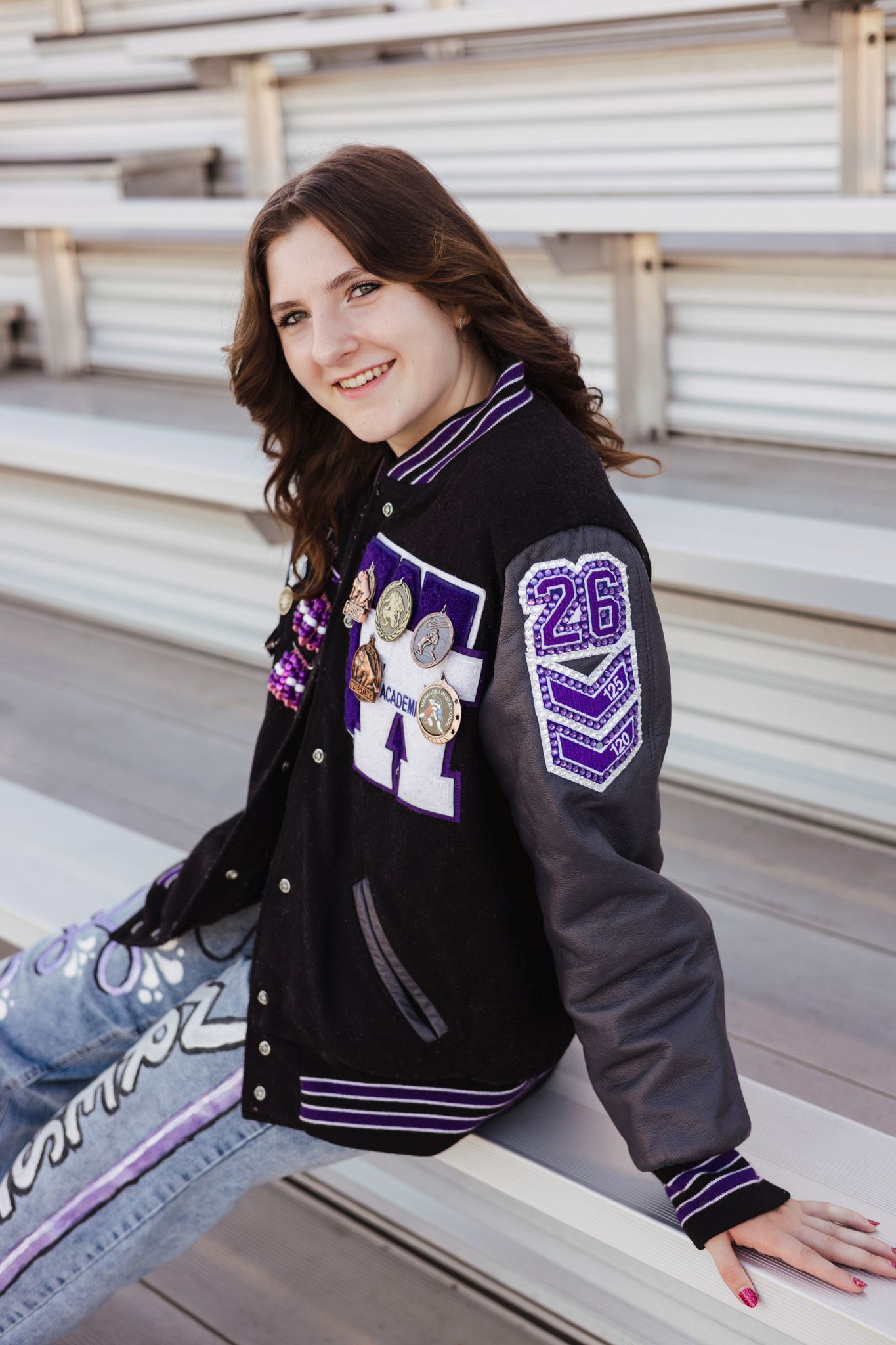 Smiling teenage girl wearing a black and purple varsity letterman jacket with patches, sitting in stadium bleachers.