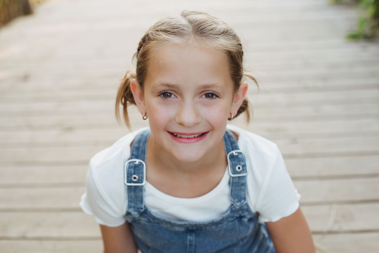 Sweet young girl in denim overalls and white shirt smiles on outdoor wooden steps.