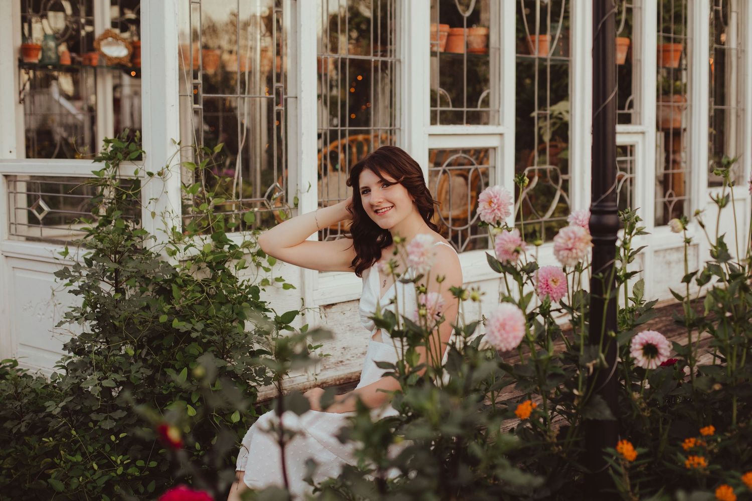 Young woman posing among colorful garden flowers near vintage white window frame.