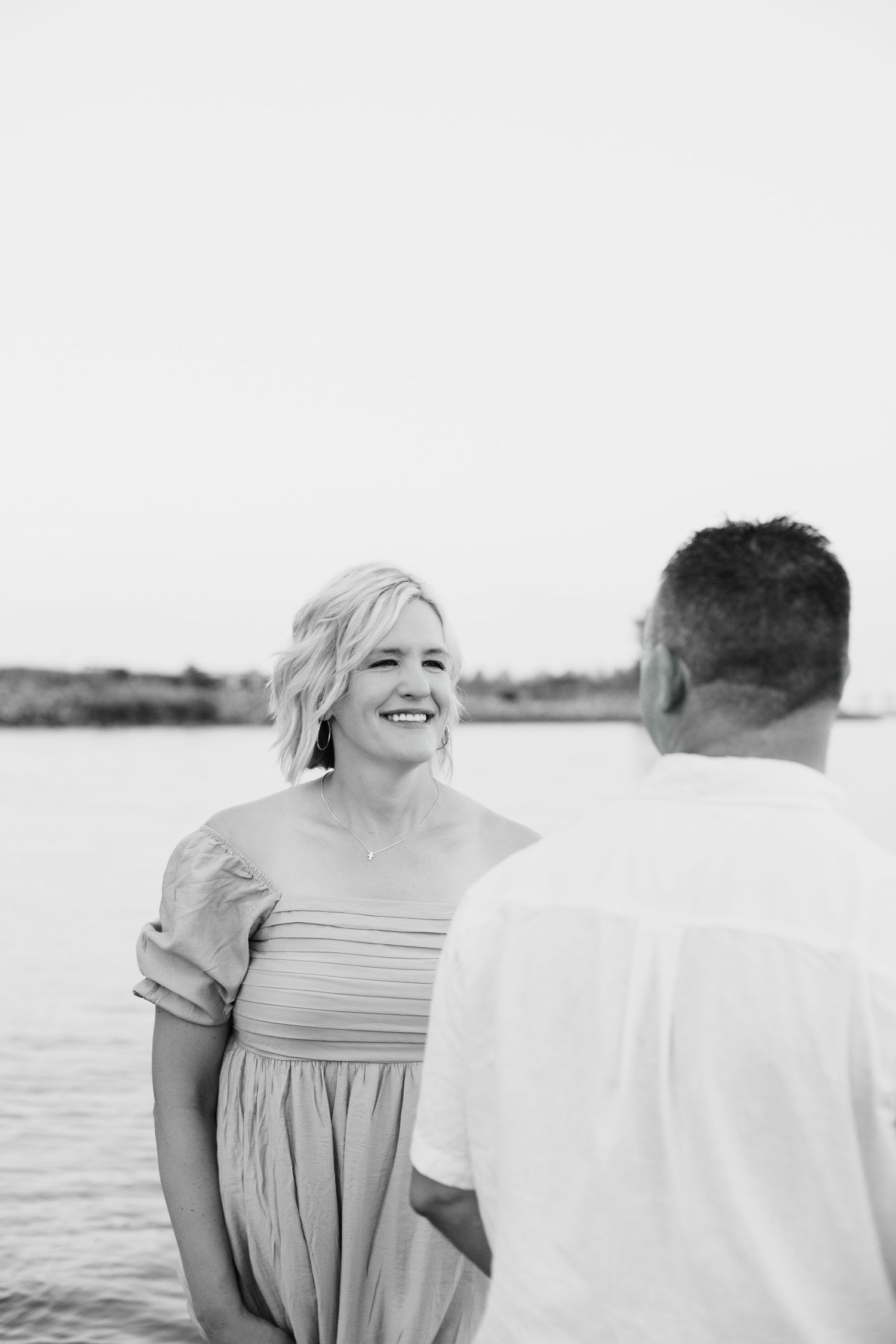 Smiling blonde woman in off-shoulder dress standing by water facing man in black and white photo.