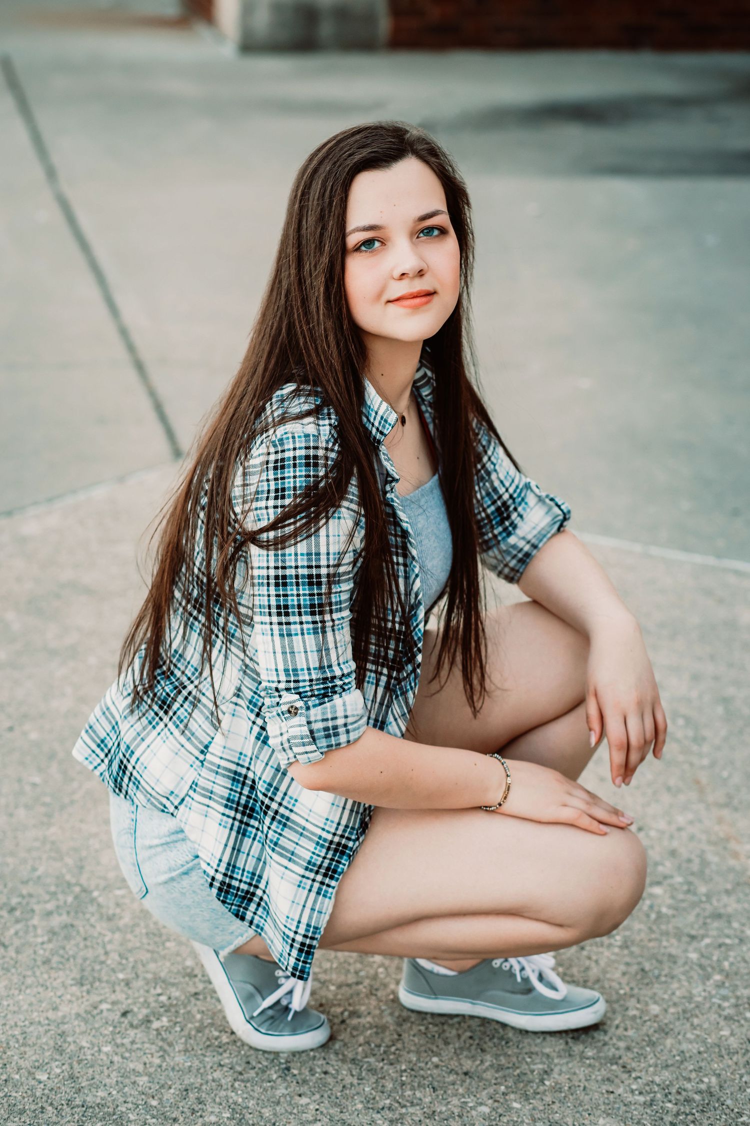 Young woman with long dark hair crouching outdoors, wearing a plaid shirt, denim shorts, and gray sneakers.