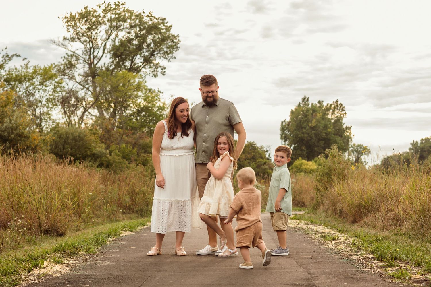 Family of five posing on a paved path surrounded by tall grass and trees during an outdoor portrait session.