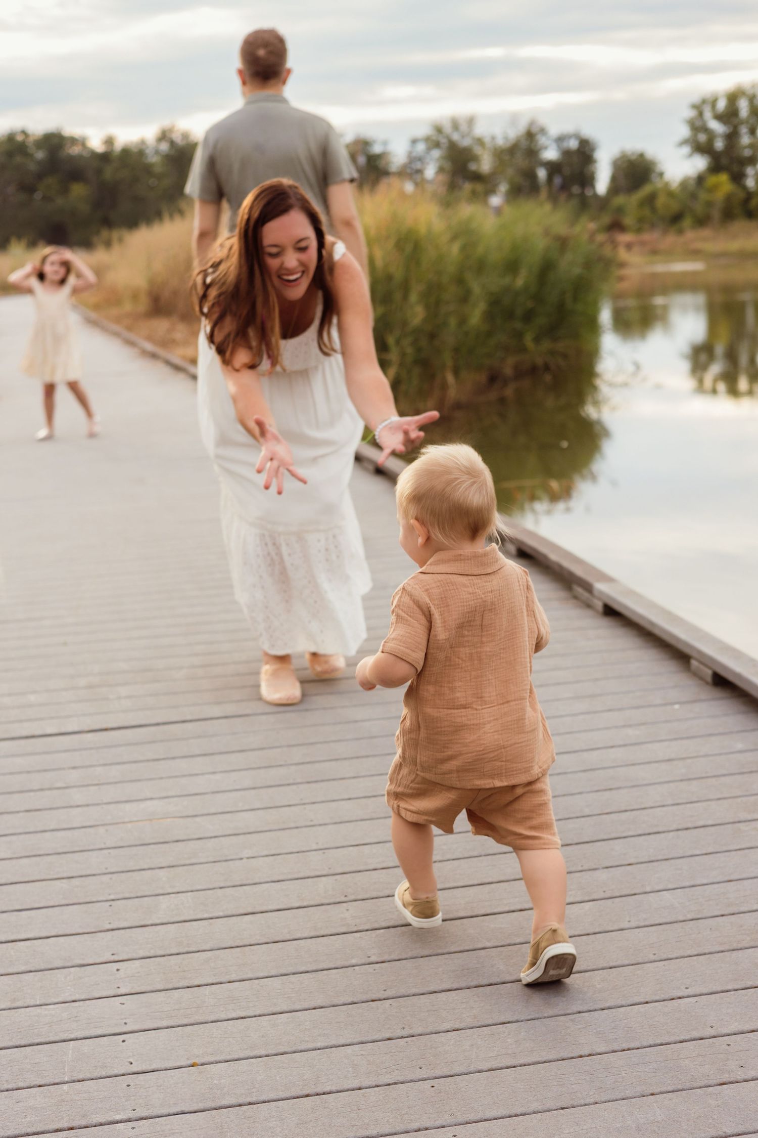 A joyful mother in a white dress reaches for her toddler on a wooden boardwalk near a lake with family nearby.