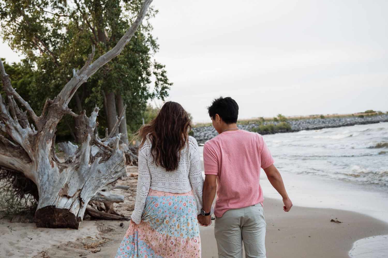 A couple walks hand in hand along a scenic beach with driftwood and ocean waves in the background.