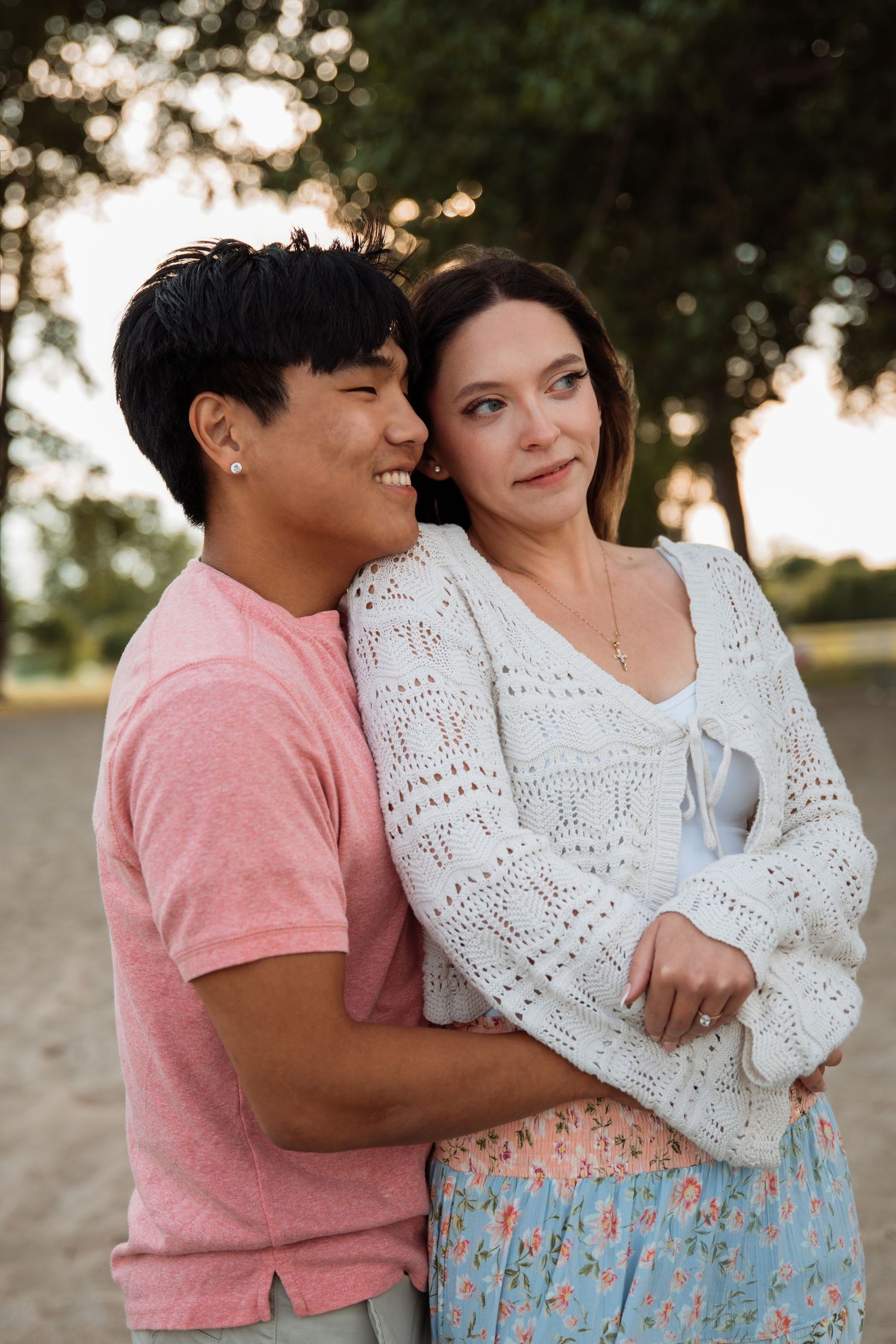 A smiling interracial couple embraces outdoors at sunset, the man in pink and woman in a white knit cardigan and floral skirt.