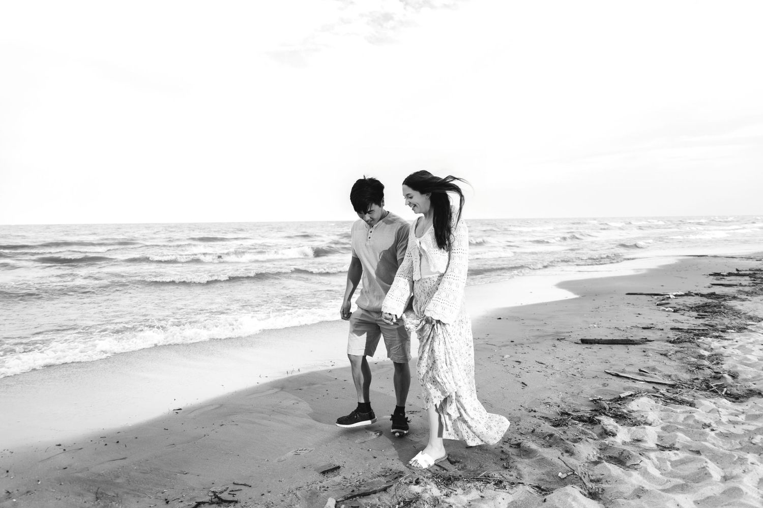 Black and white photo of a couple walking hand in hand along a beach shoreline with ocean waves.
