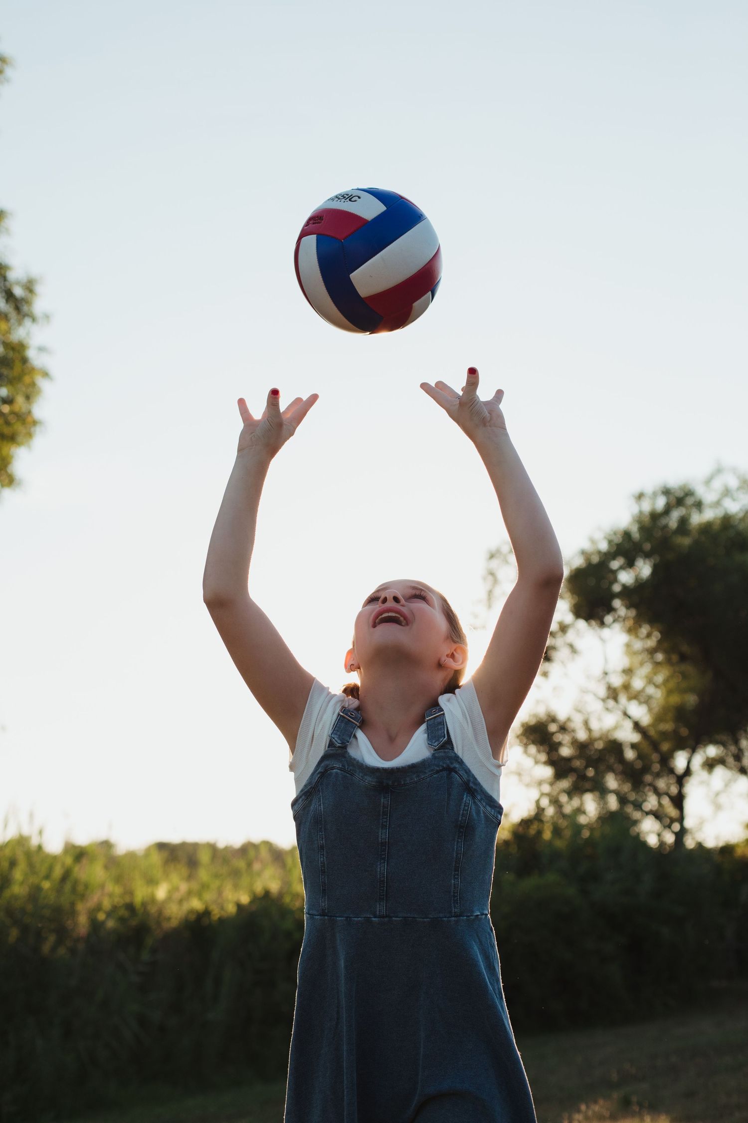 Young girl in denim overalls tossing a colorful volleyball into the air outdoors.