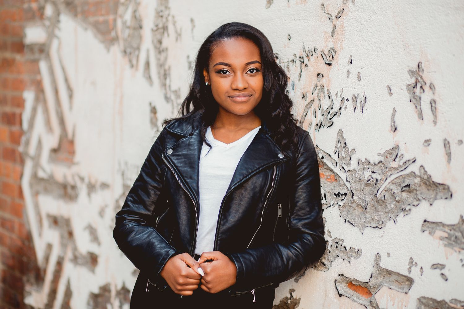 Young girl in black leather jacket poses confidently against a rustic peeling white painted brick wall.