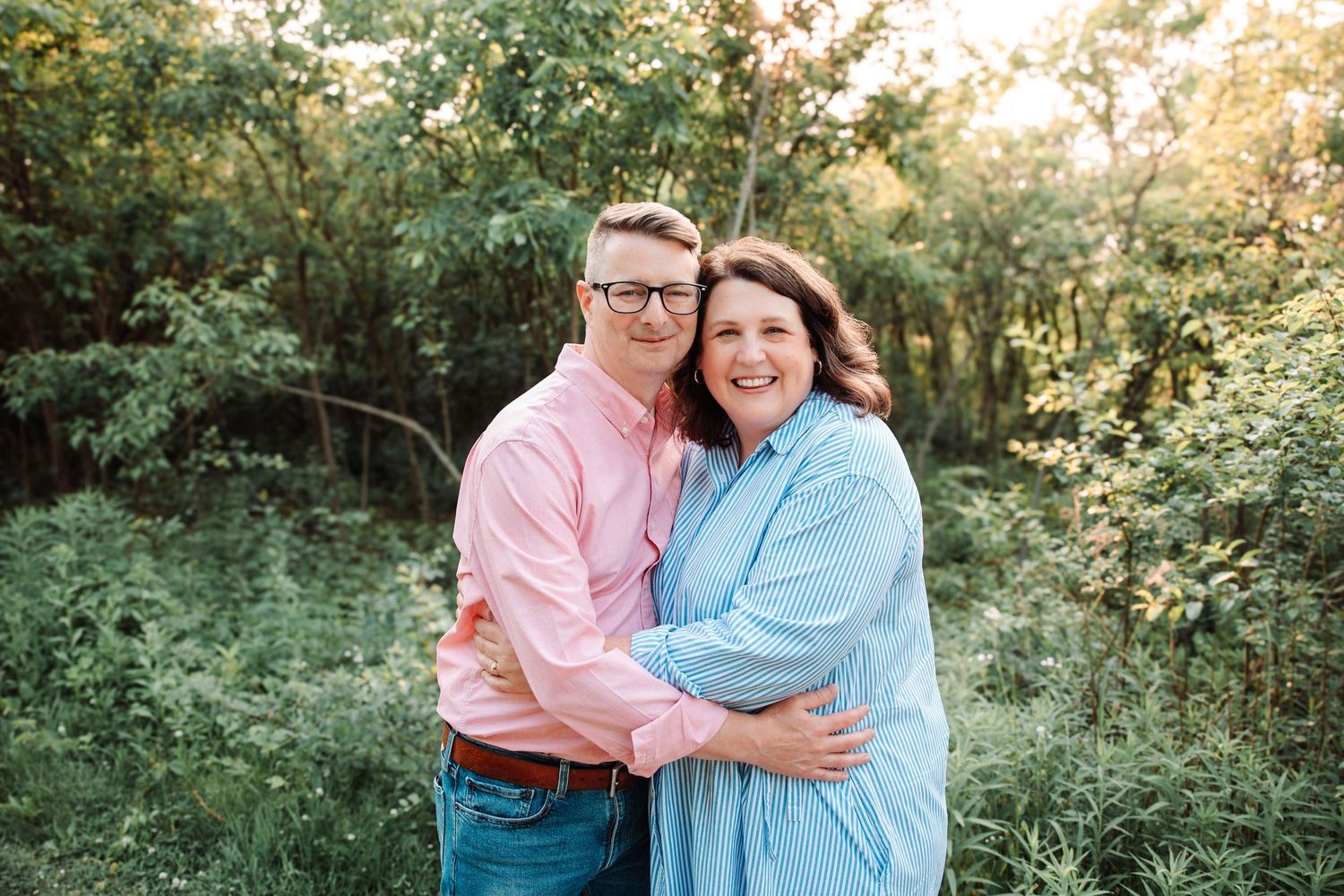 Couple embracing and smiling outdoors surrounded by lush green trees and golden sunlight.