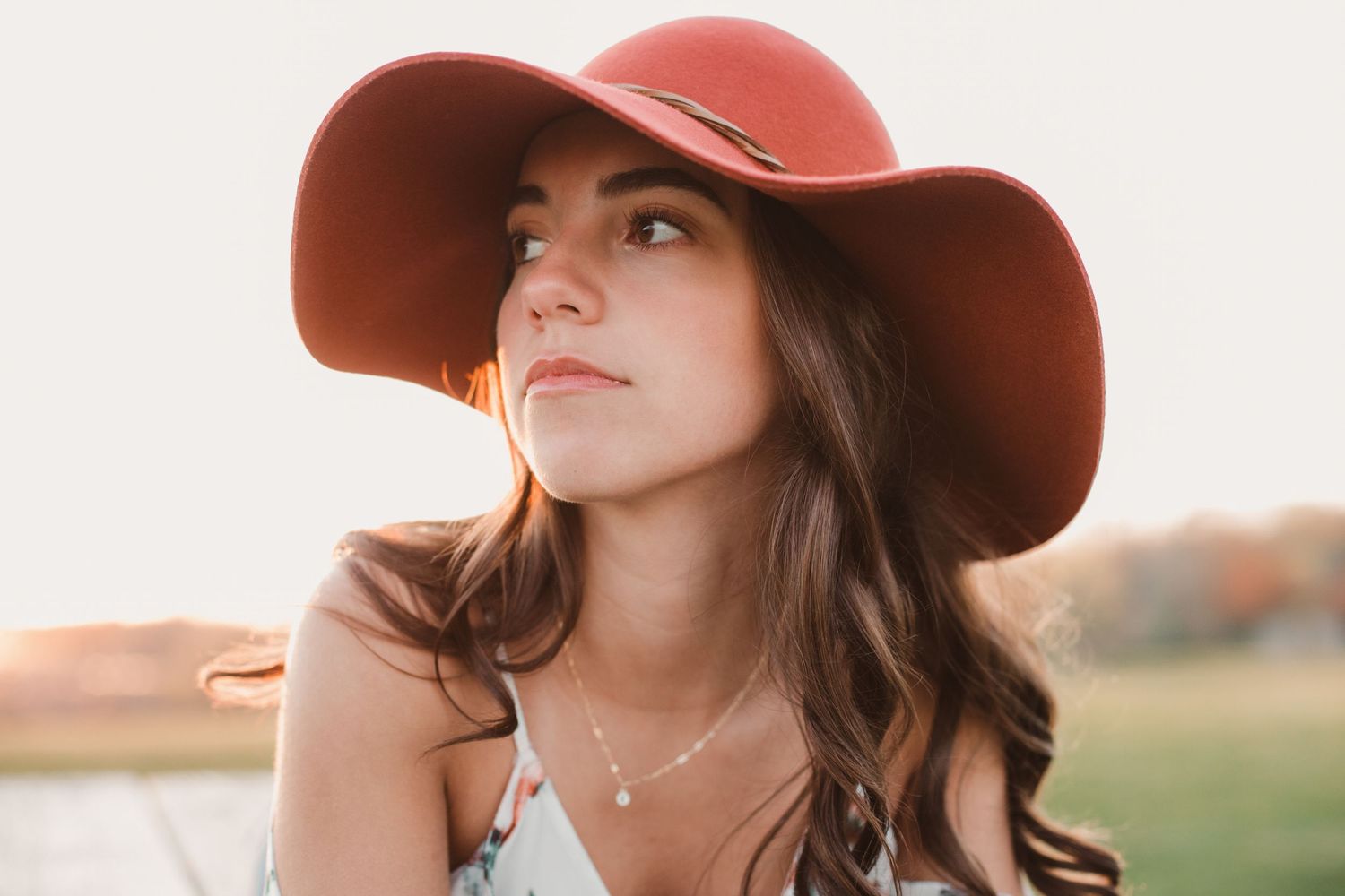 Beautiful brunette woman wearing a wide-brim rust-colored hat gazing into the distance at golden sunset.