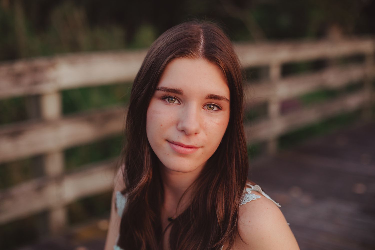 Close-up portrait of brunette teen girl near wooden fence in warm natural outdoor lighting.