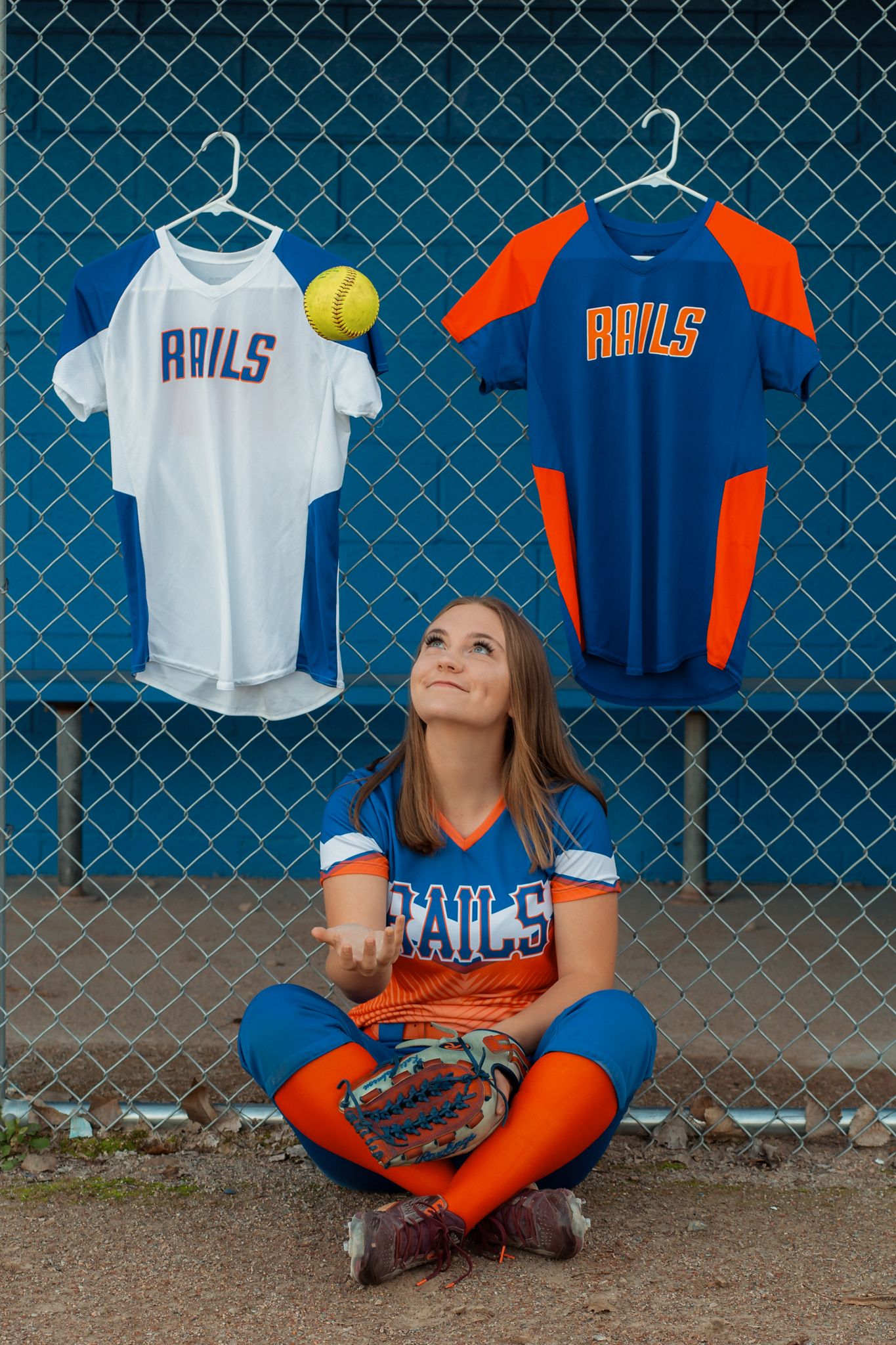 Softball player in Rails uniform sitting by a fence tossing a ball with jerseys hanging behind her.