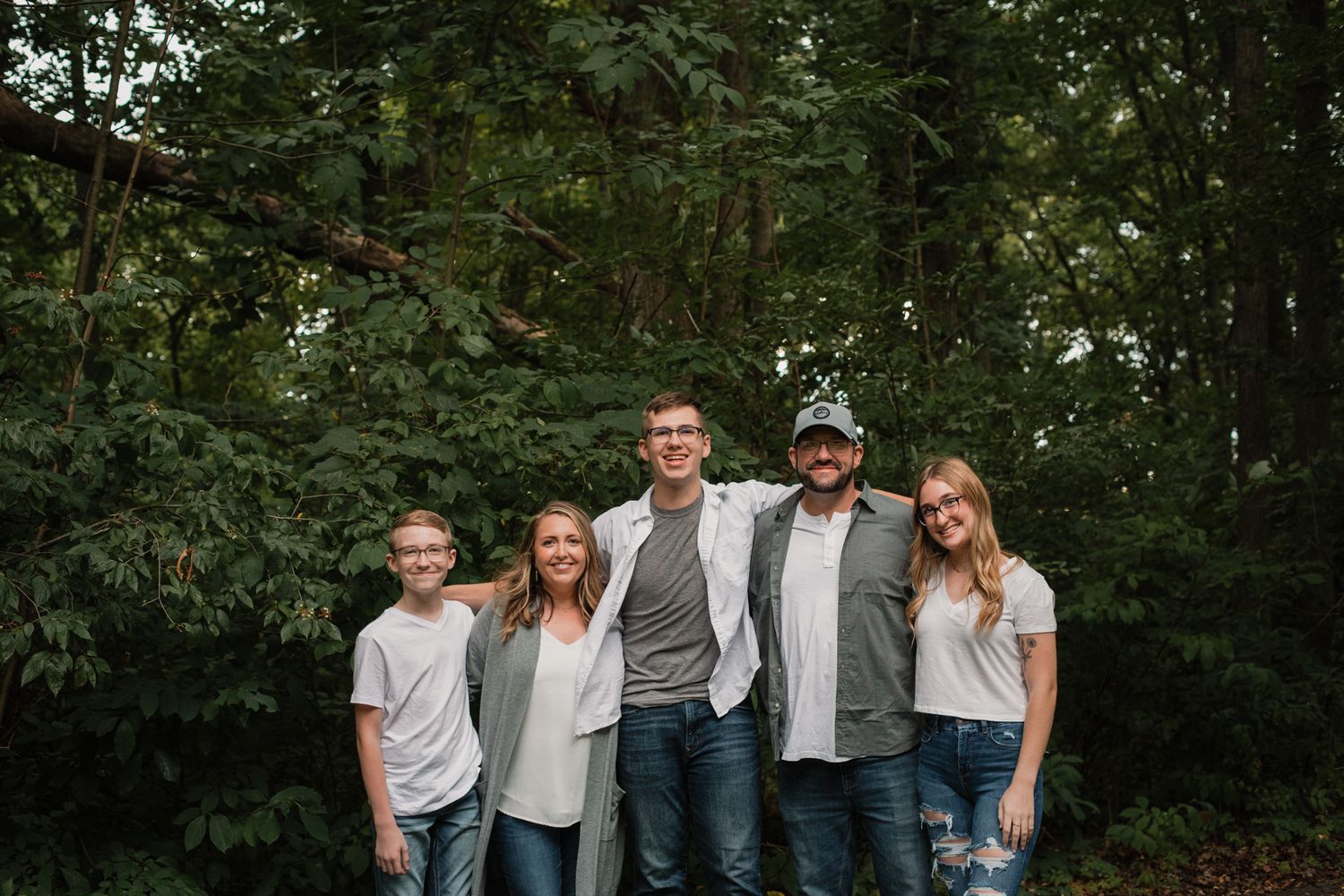 A happy family of five stands together outdoors surrounded by green trees in matching casual outfits.