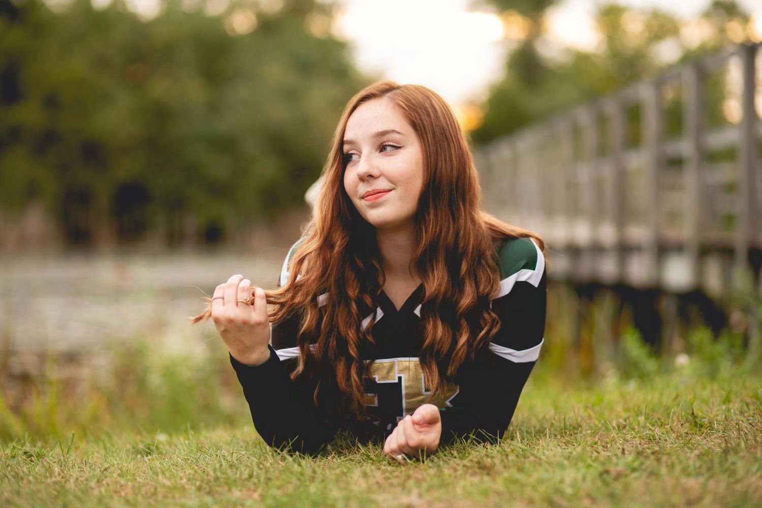 A young redhead woman in a sports jersey lies on green grass, smiling, holding a letter, with a wooden bridge behind her.