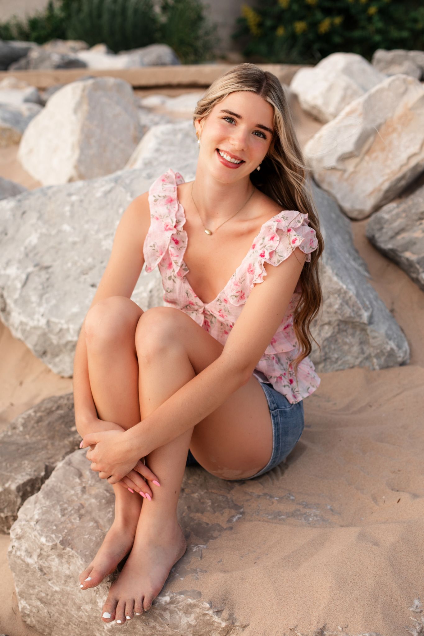 Young woman in floral top and denim shorts sitting barefoot on sandy rocks, smiling outdoors.
